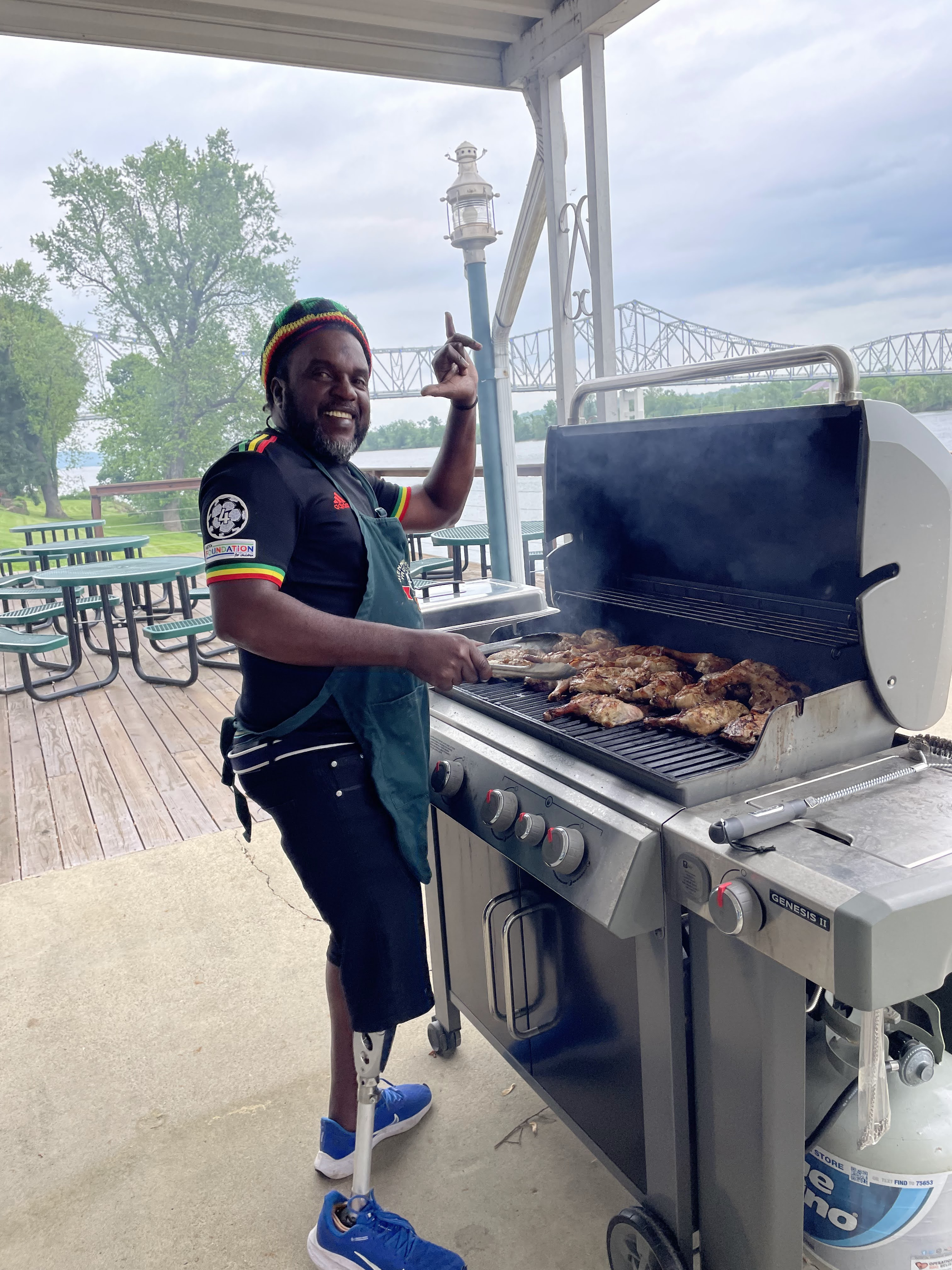 A man with a prosthetic leg grilling chicken on an outdoor barbecue grill near a river, with picnic tables and trees in the background.