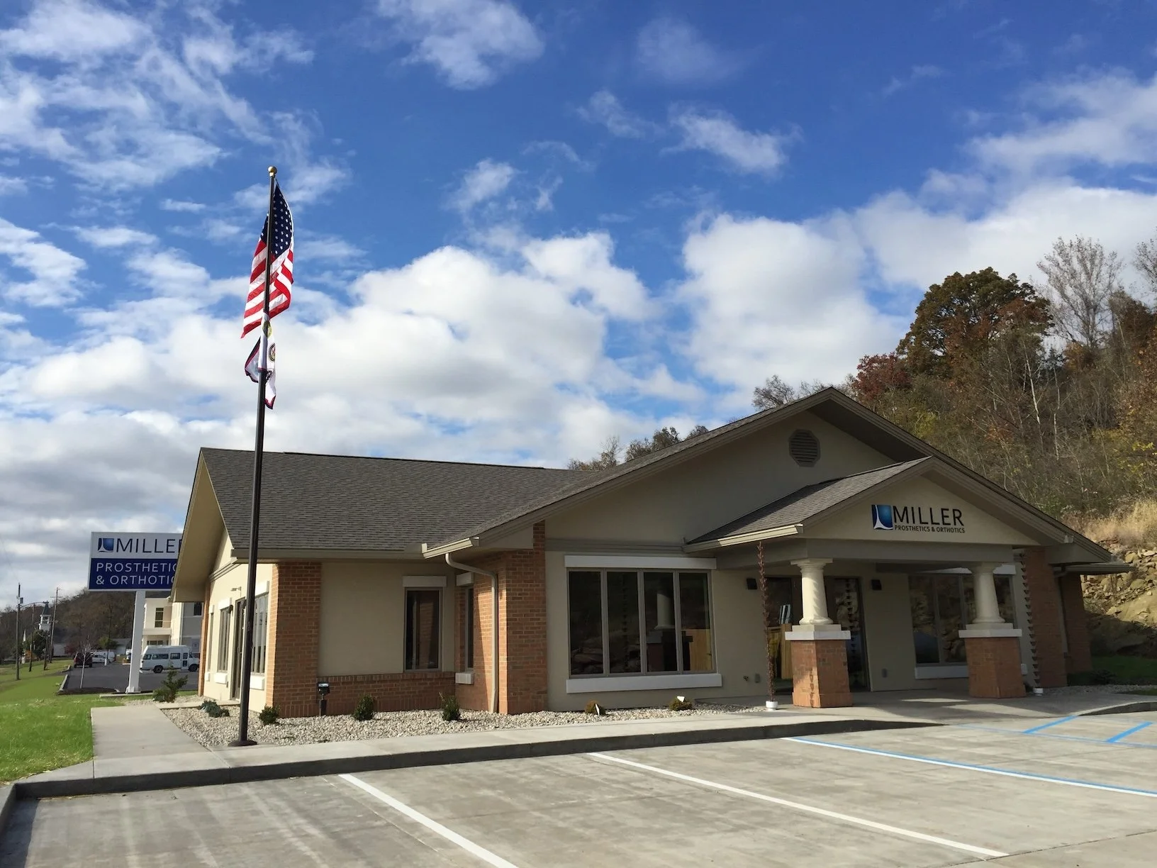 A one-story building with a sign reading 'MILLER PROSTHETICS & ORTHOTICS' and flags flying outside, situated in a parking lot with a clear sky and autumn trees in the background.