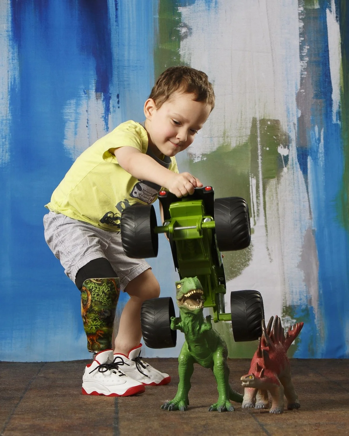 A young boy playing with a green toy monster truck and dinosaur figurines on a brown carpeted floor, with a colorful painted backdrop.