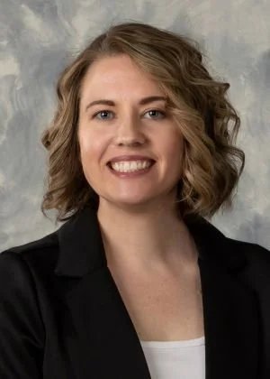 A woman with shoulder-length wavy hair, smiling, wearing a black blazer over a white top, against a gray textured background.