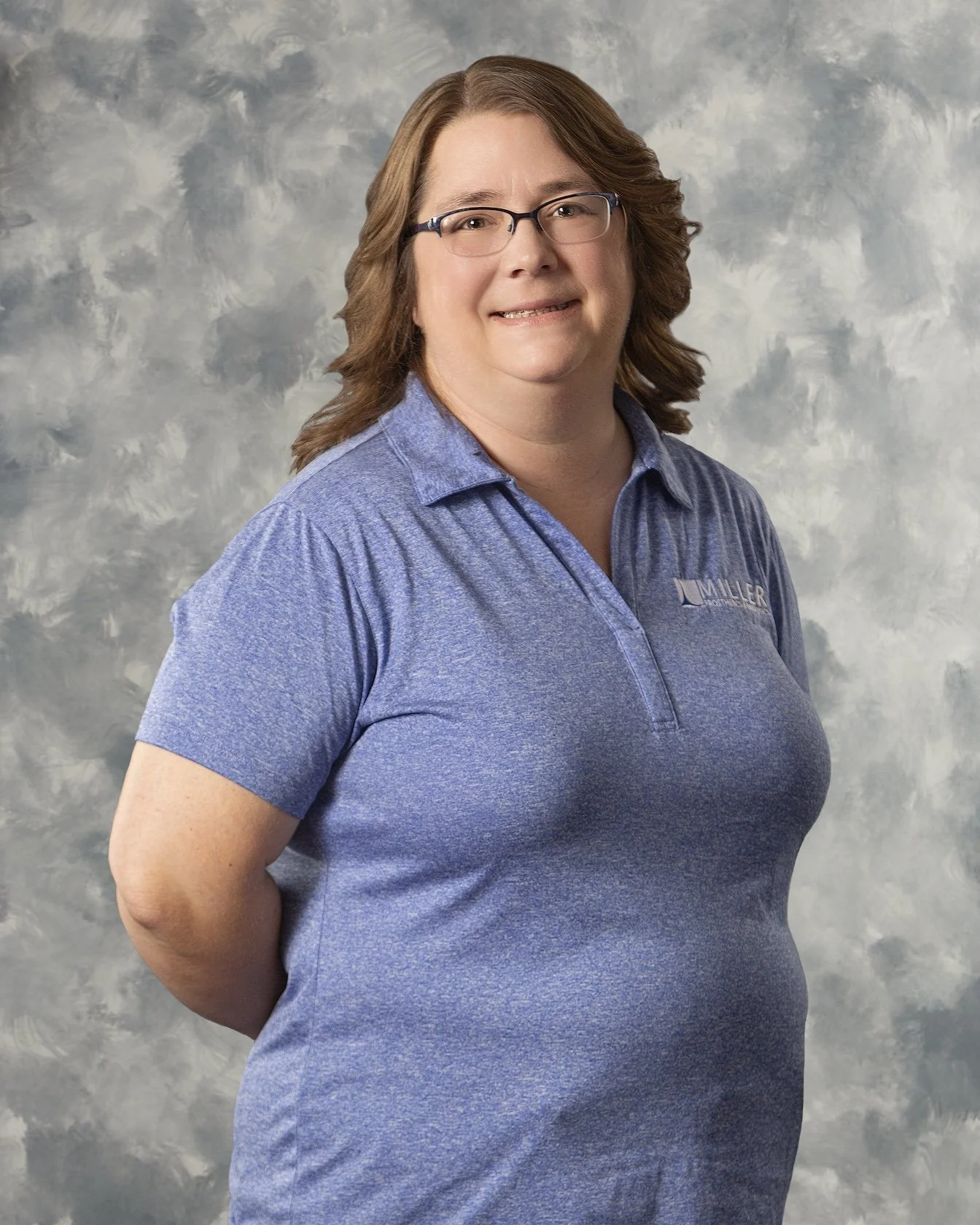 A woman with shoulder-length brown hair, glasses, and a blue collared shirt with a logo, standing in front of a cloudy gray background.