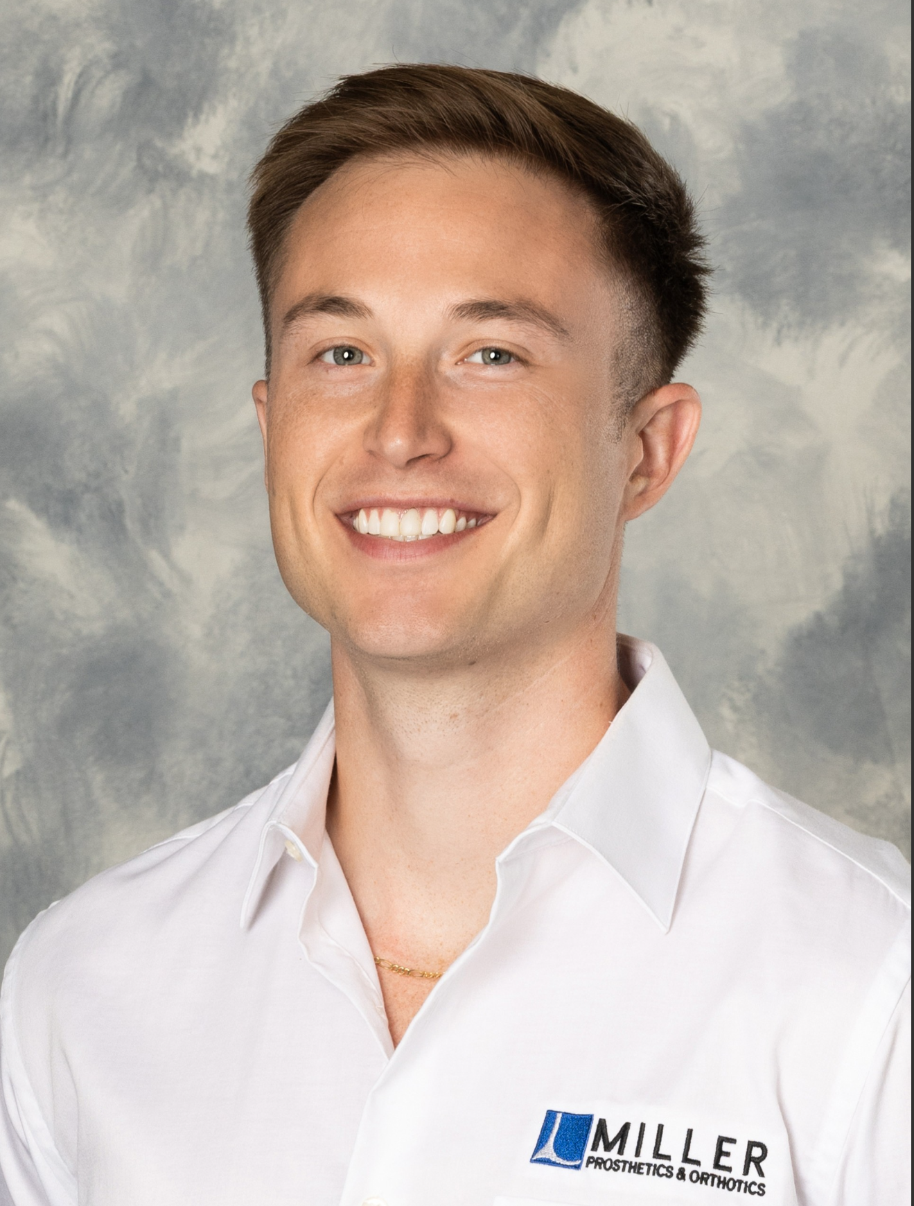 Portrait of a young man with short brown hair, wearing a white collared shirt with a logo for Miller Prosthetics & Orthotics, smiling against a textured background.