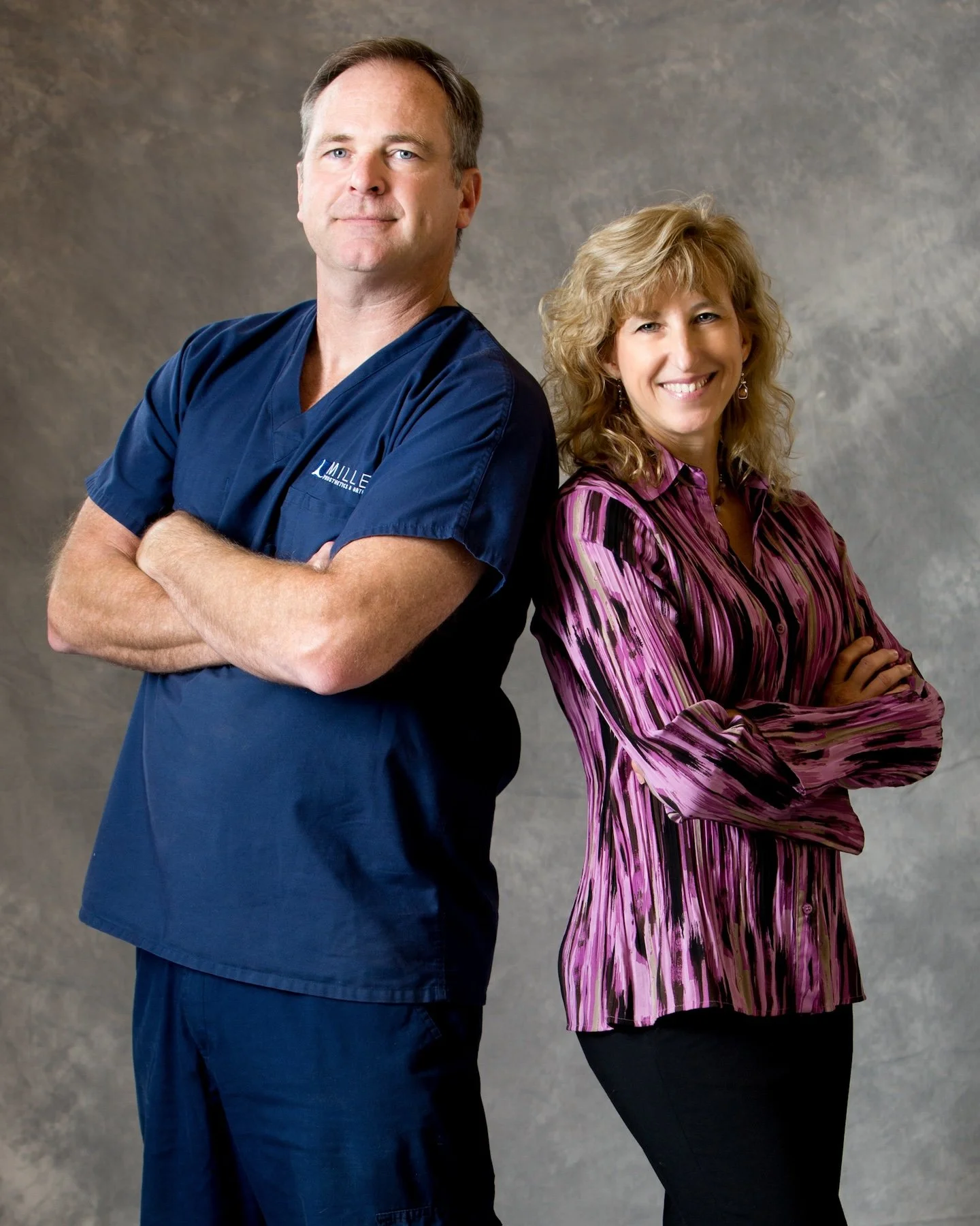 A man in blue medical scrubs and a woman in a pink and purple striped blouse pose with arms crossed against a gray background. The man is standing in front with his arms folded, and the woman is standing slightly behind him, also with her arms crossed, smiling at the camera.