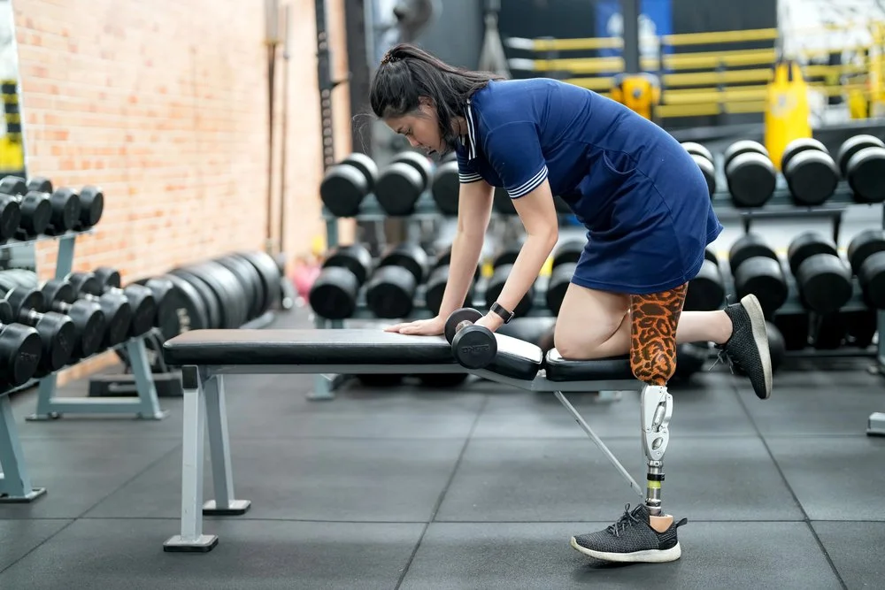 A woman with a prosthetic leg working out in a gym, performing a exercise with dumbbells on a workout bench.