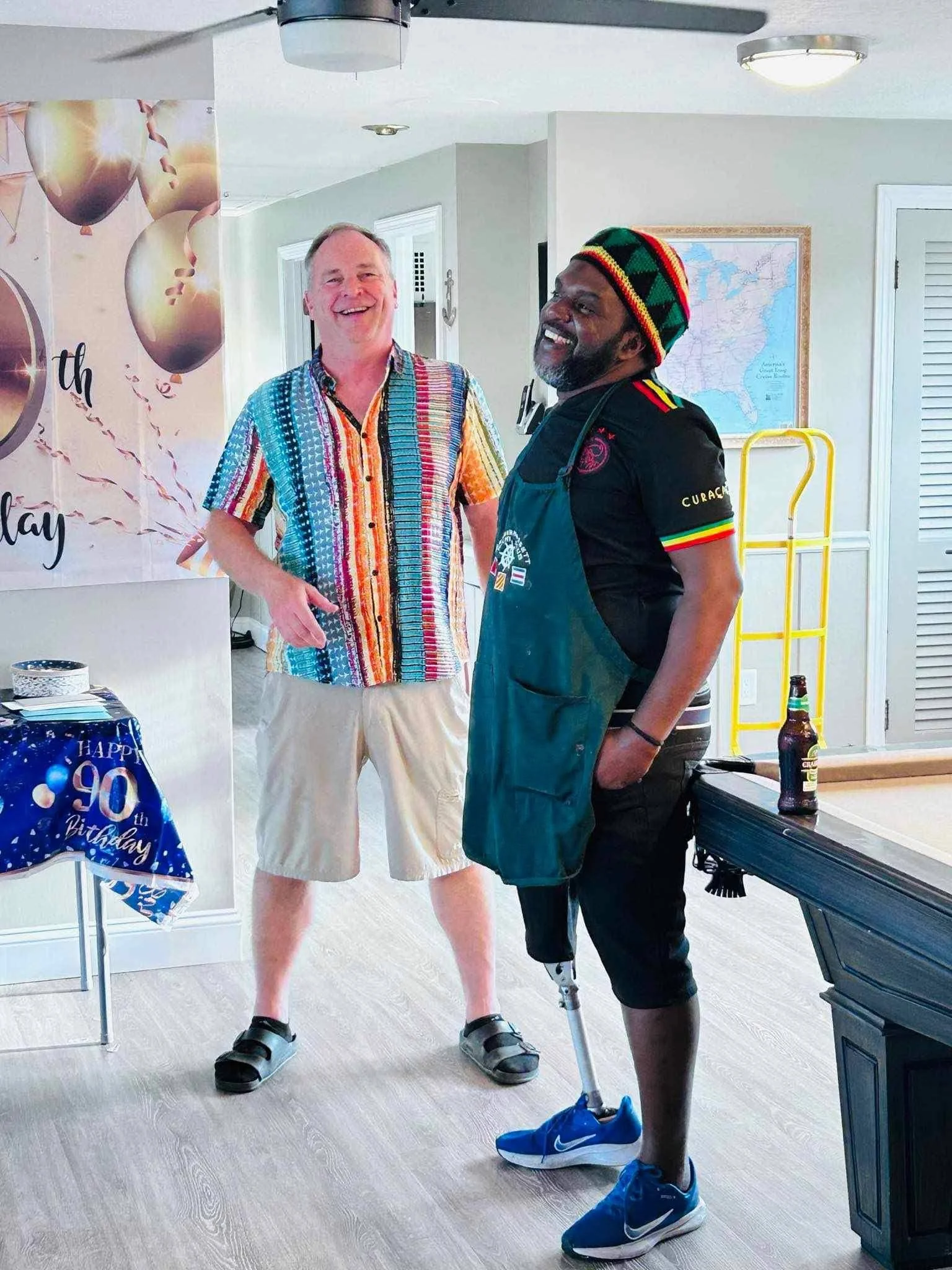 Two men are standing and smiling. One is wearing a colorful, striped shirt and beige shorts, and the other has a prosthetic leg, a black shirt with rainbow-colored accents, and a multicolored knit cap. There is a birthday banner and a table with a 'H