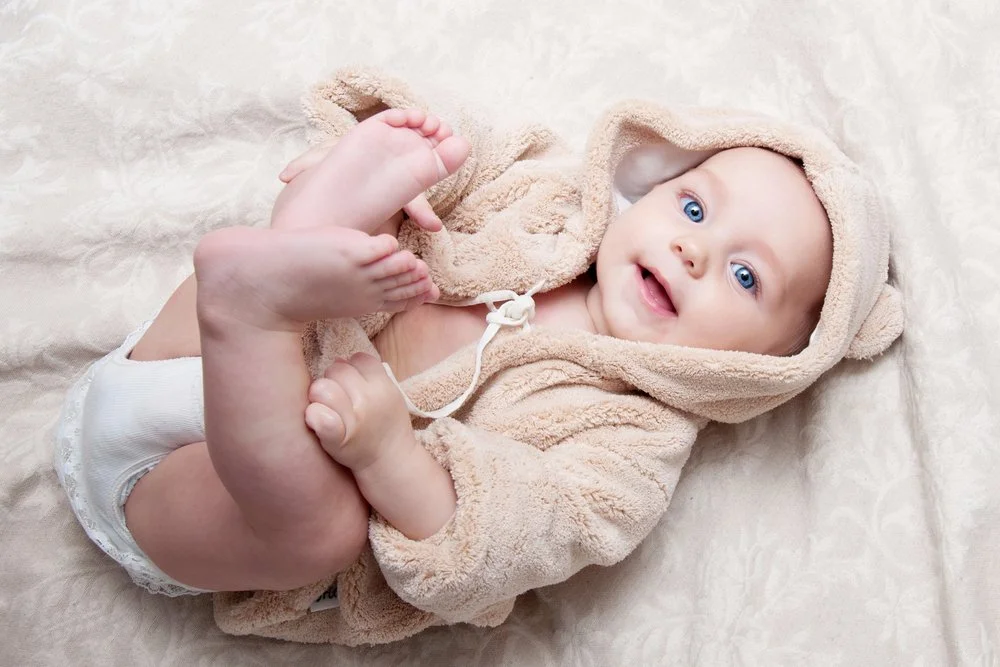 Adorable baby with blue eyes lying on a soft surface, wearing a beige hooded fuzzy jacket with bear ears, and a diaper, holding its feet.
