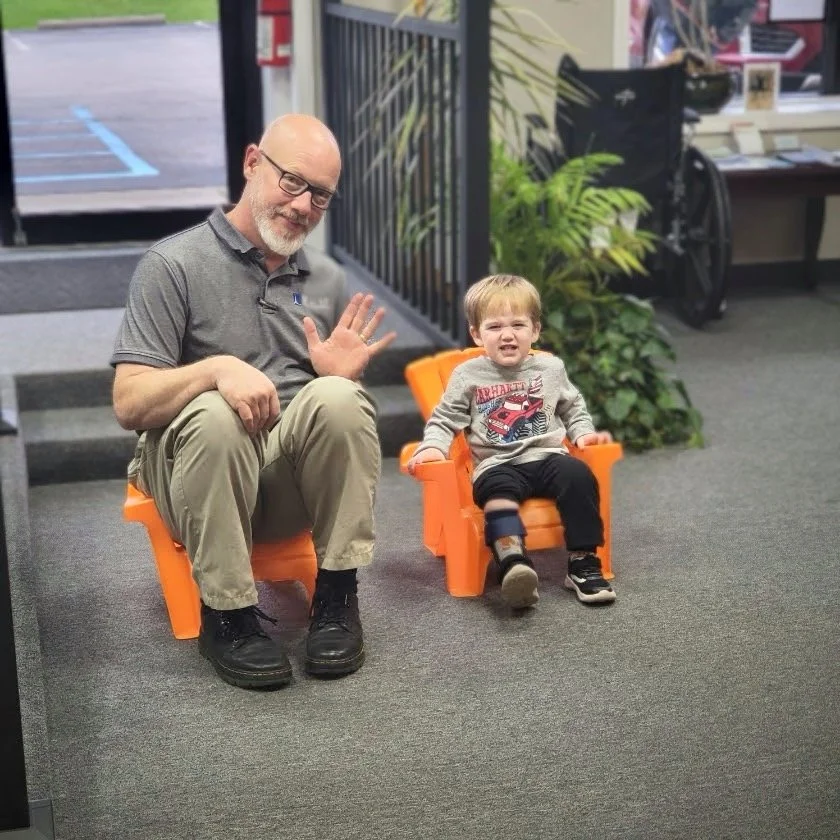 An older man and a young boy sitting on small orange chairs indoors, with the man waving and smiling, and the boy looking distressed.