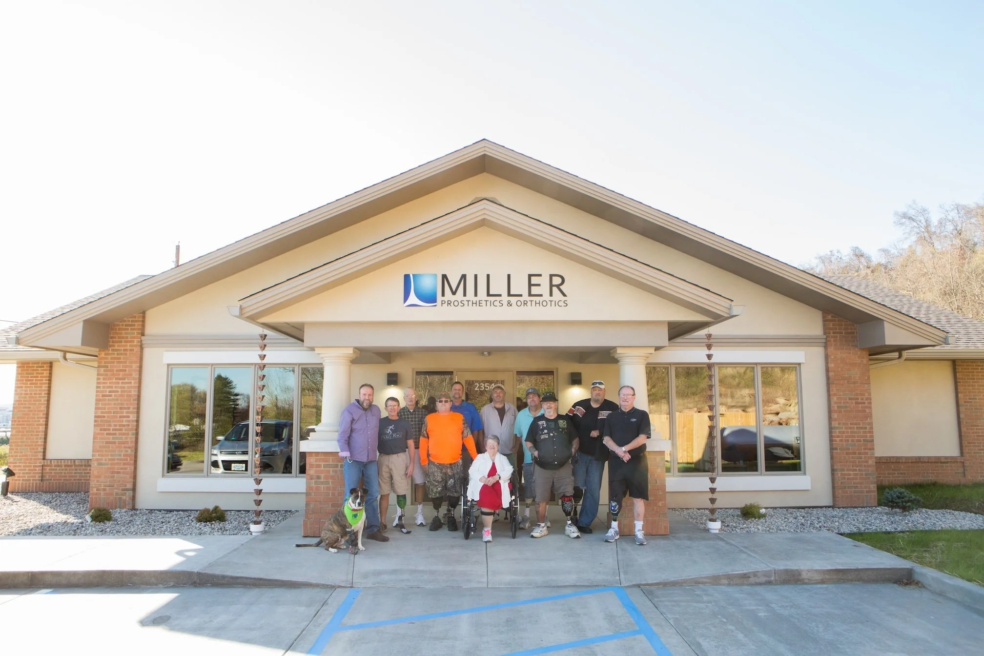 Group of people, including a young girl in a wheelchair and a service dog, standing in front of Miller Prosthetics & Orthotics building on a sunny day.