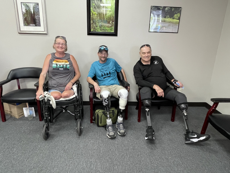 Three people sitting on chairs in a room, two with prosthetic legs, smiling at the camera.