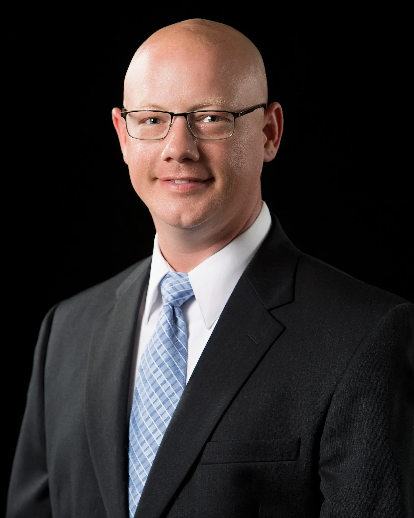 Professional headshot of a bald man wearing glasses, a black suit, white shirt, and light blue tie against a black background.