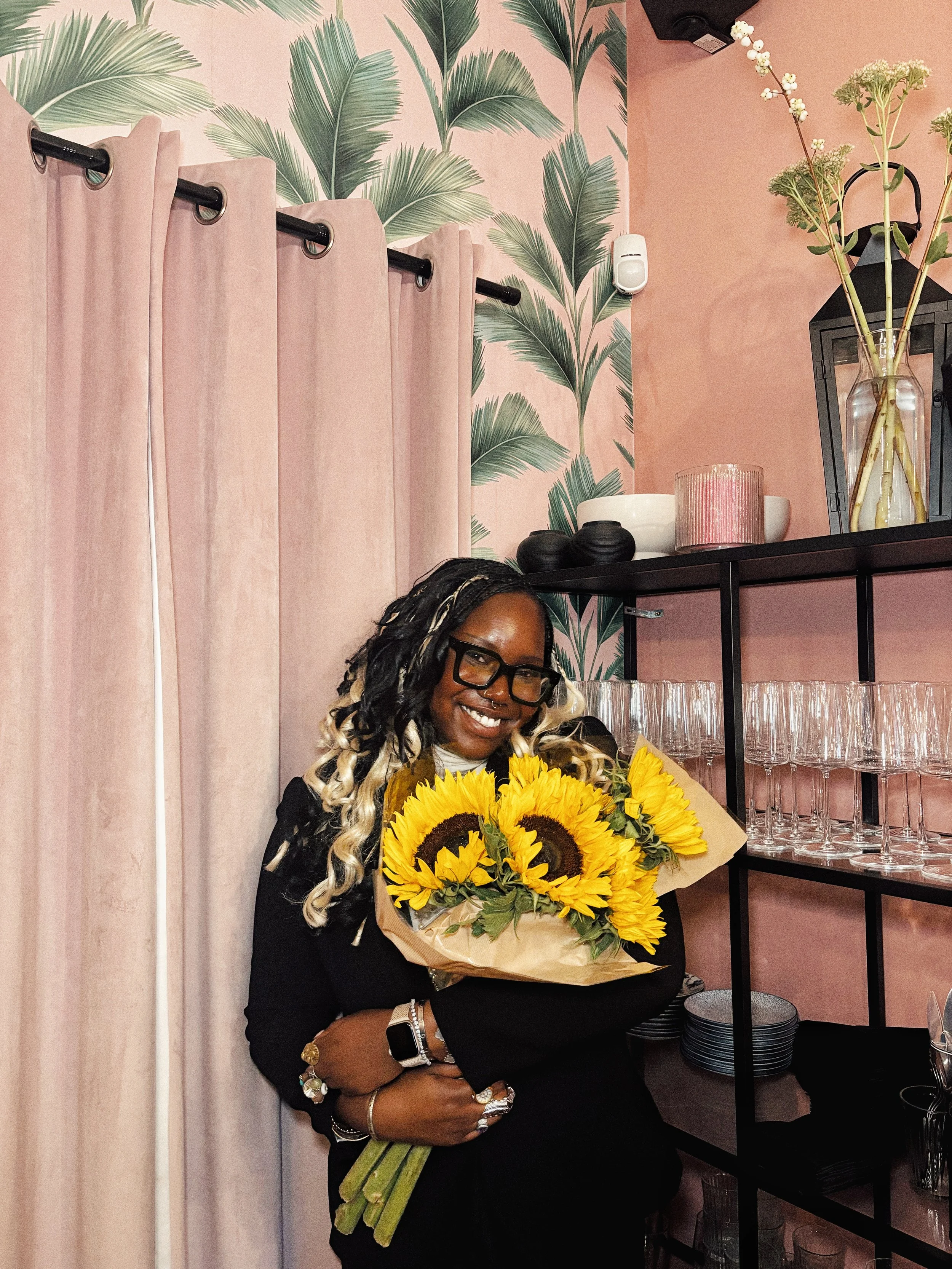 A woman smiling and holding a bouquet of sunflowers in a room with pink curtains, palm leaf wallpaper, and glassware on shelves.