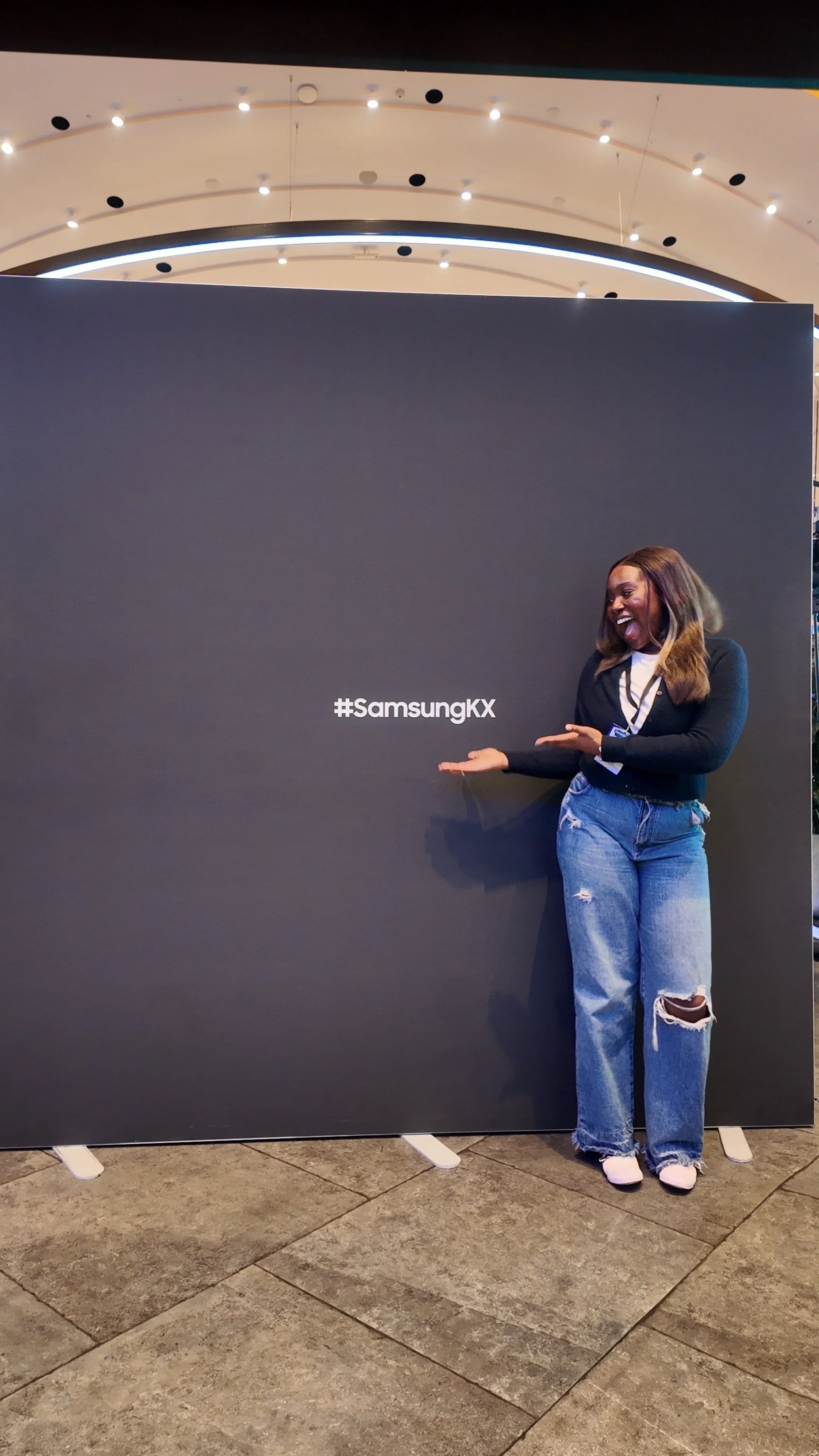 A woman standing next to a large black display with the hashtag #SamsungKX written on it, smiling and pointing towards the hashtag.