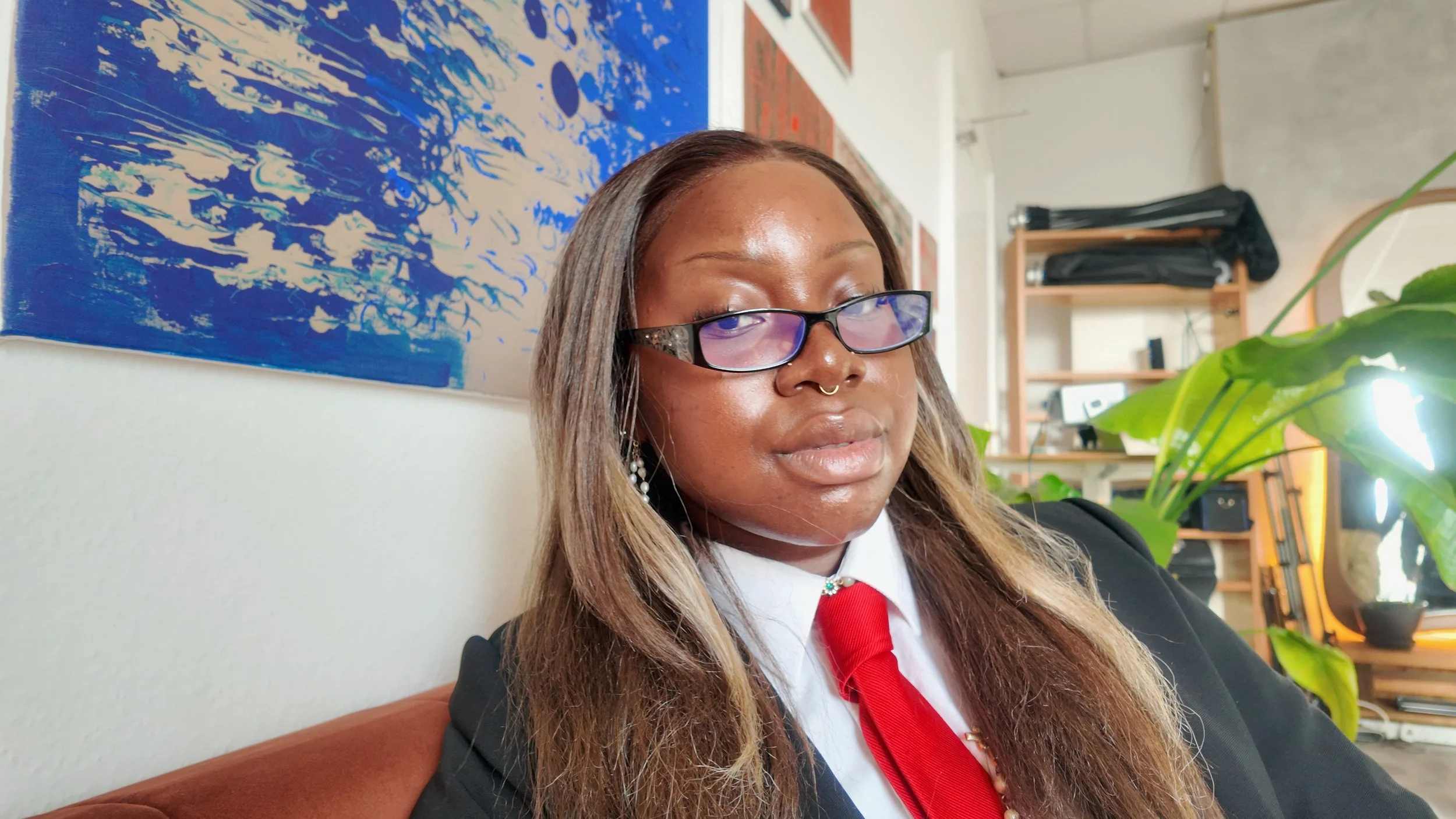 A woman with long blonde hair, glasses, and a septum piercing sitting on a beige couch, wearing a black blazer, white shirt, and red tie. Behind her is a large blue and white abstract painting, some green houseplants, and a wooden shelf with black bags and electronic equipment. Natural light is coming from the right side.