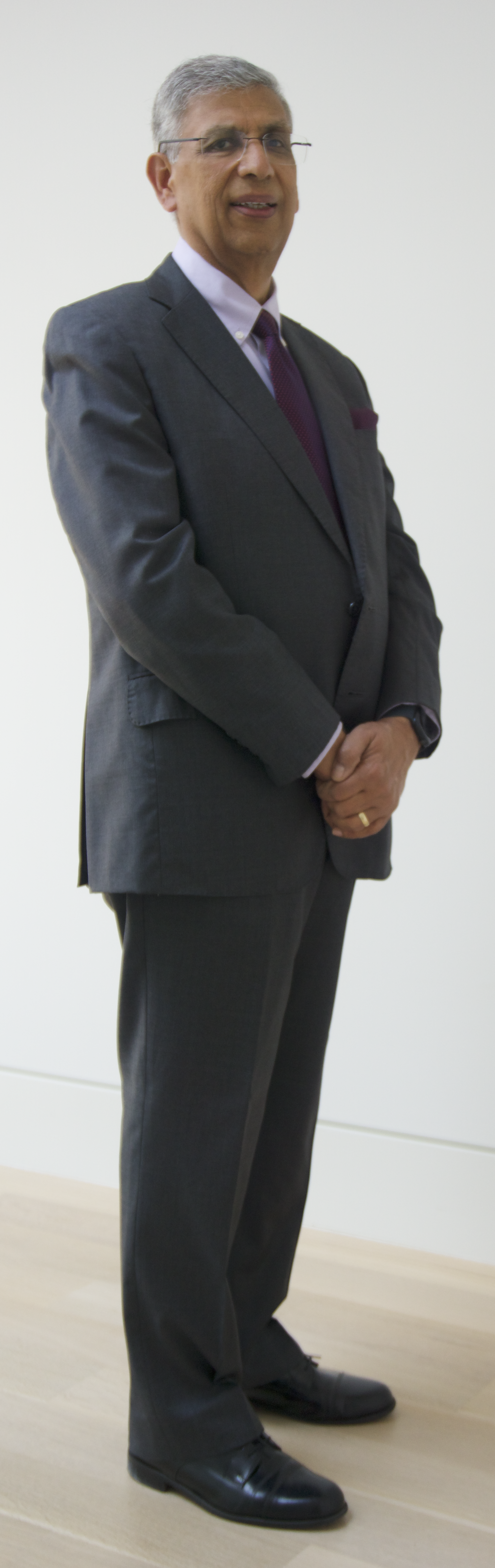 Man in a dark gray business suit, white shirt, purple tie, wearing glasses, standing with hands clasped, in front of a plain white wall.