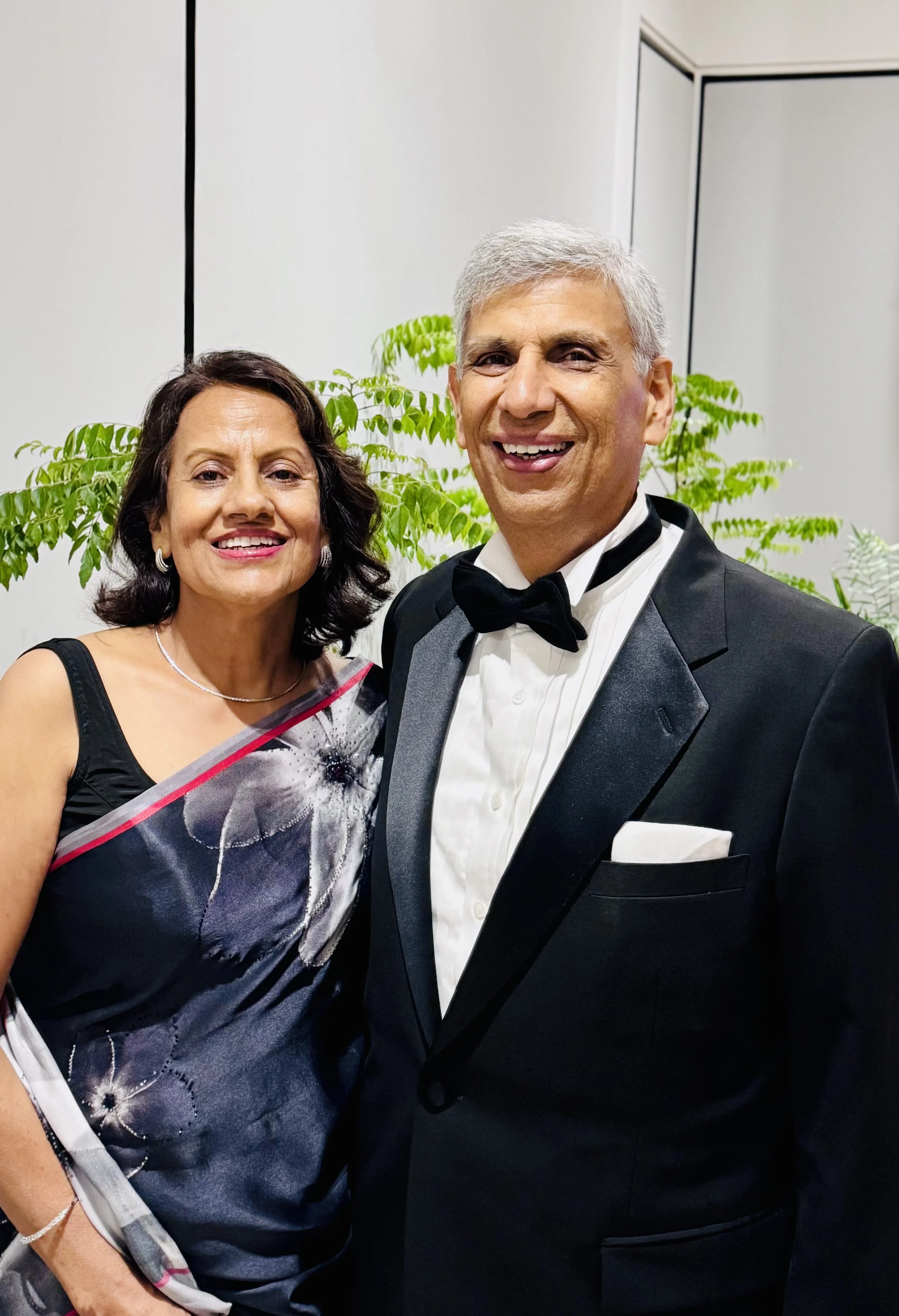 A smiling middle-aged woman in a black and white dress with floral patterns, and a smiling older man in a tuxedo with a bow tie, posing together in front of green plants.