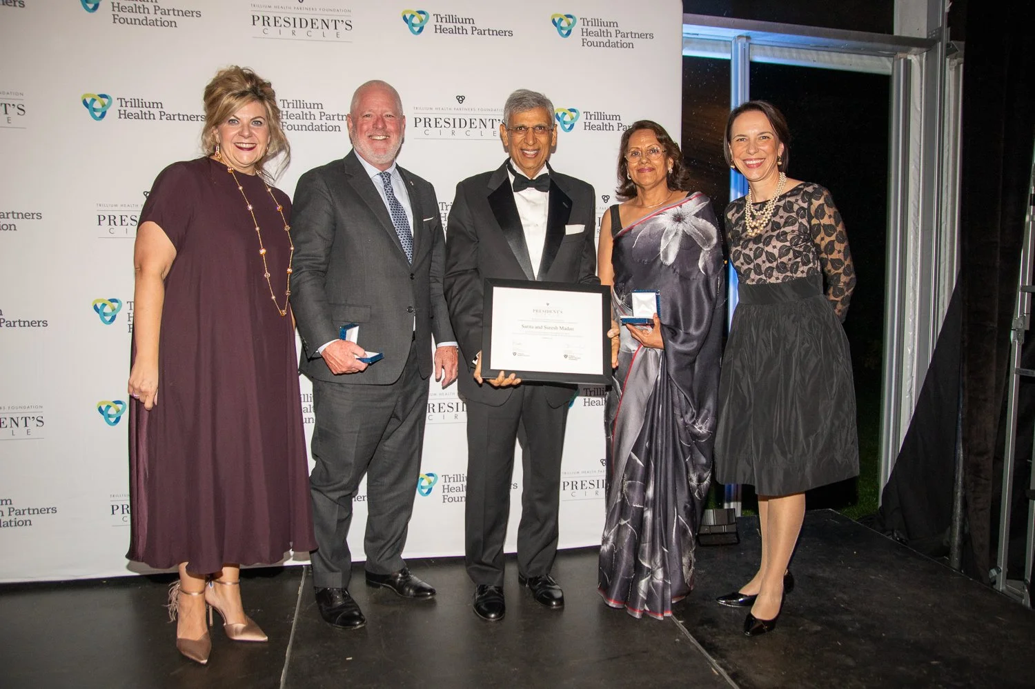 Group of six people standing on stage at an award ceremony, holding medals and a framed certificate, with a backdrop displaying 'Trillium Health Partners Foundation' and 'President's Circle' logos.