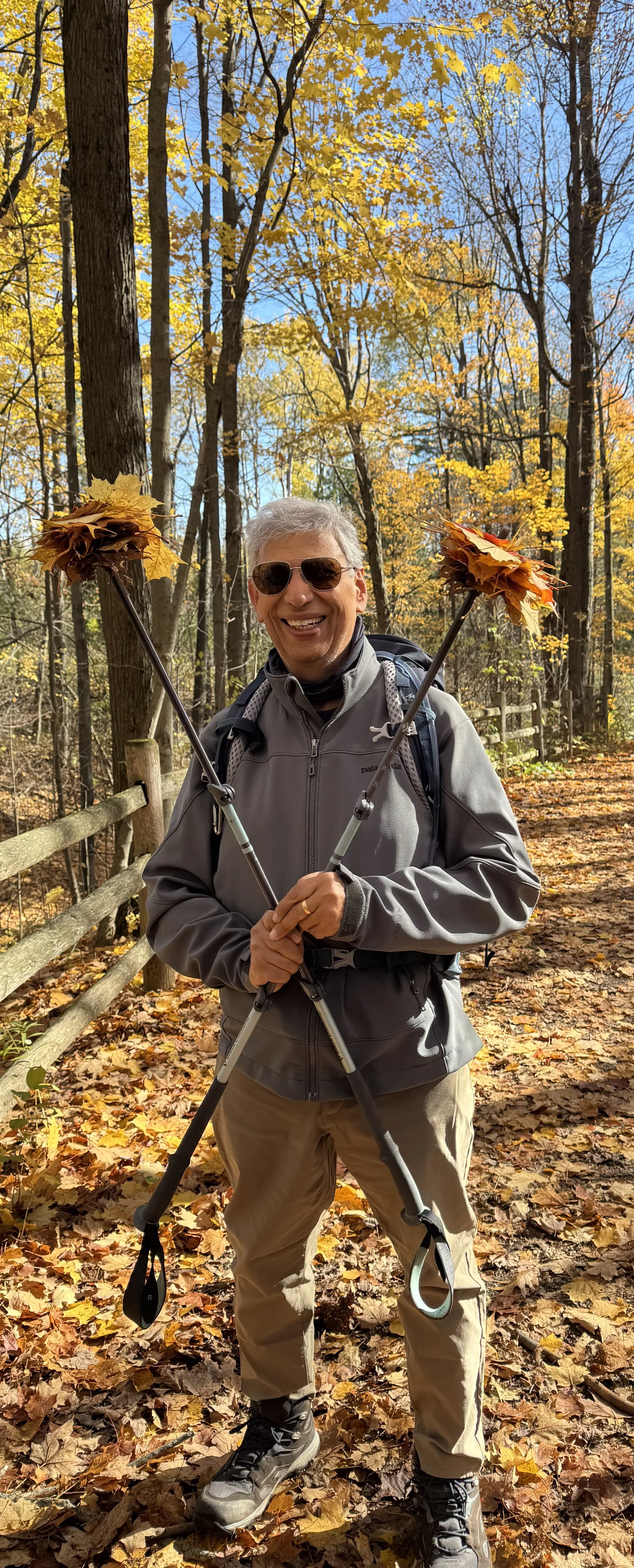 A smiling man with gray hair and sunglasses standing outdoors in a fall forest, holding a walking stick with autumn leaves.