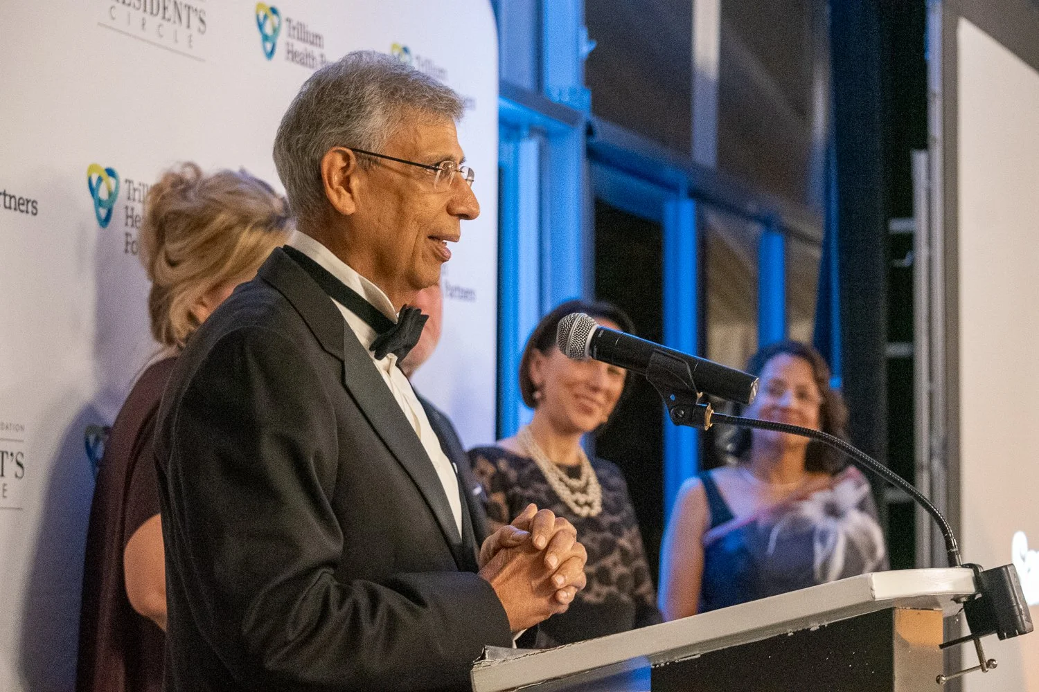A man in a tuxedo with a bowtie speaking at a podium during an event, with women standing behind him, smiling, and a backdrop displaying a logo and text.