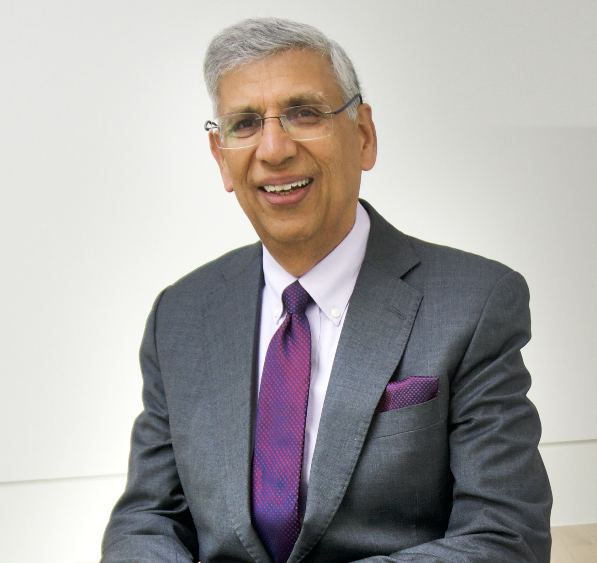 Man with gray hair, glasses, and a suit sitting in an office, smiling.