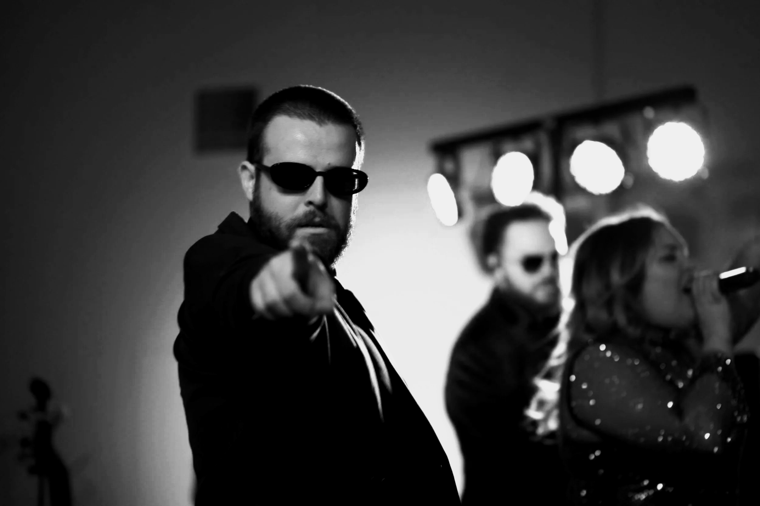 Black and white photo of three musicians with dark sunglasses, with the man in the foreground pointing towards the camera, on stage with bright lights behind them.