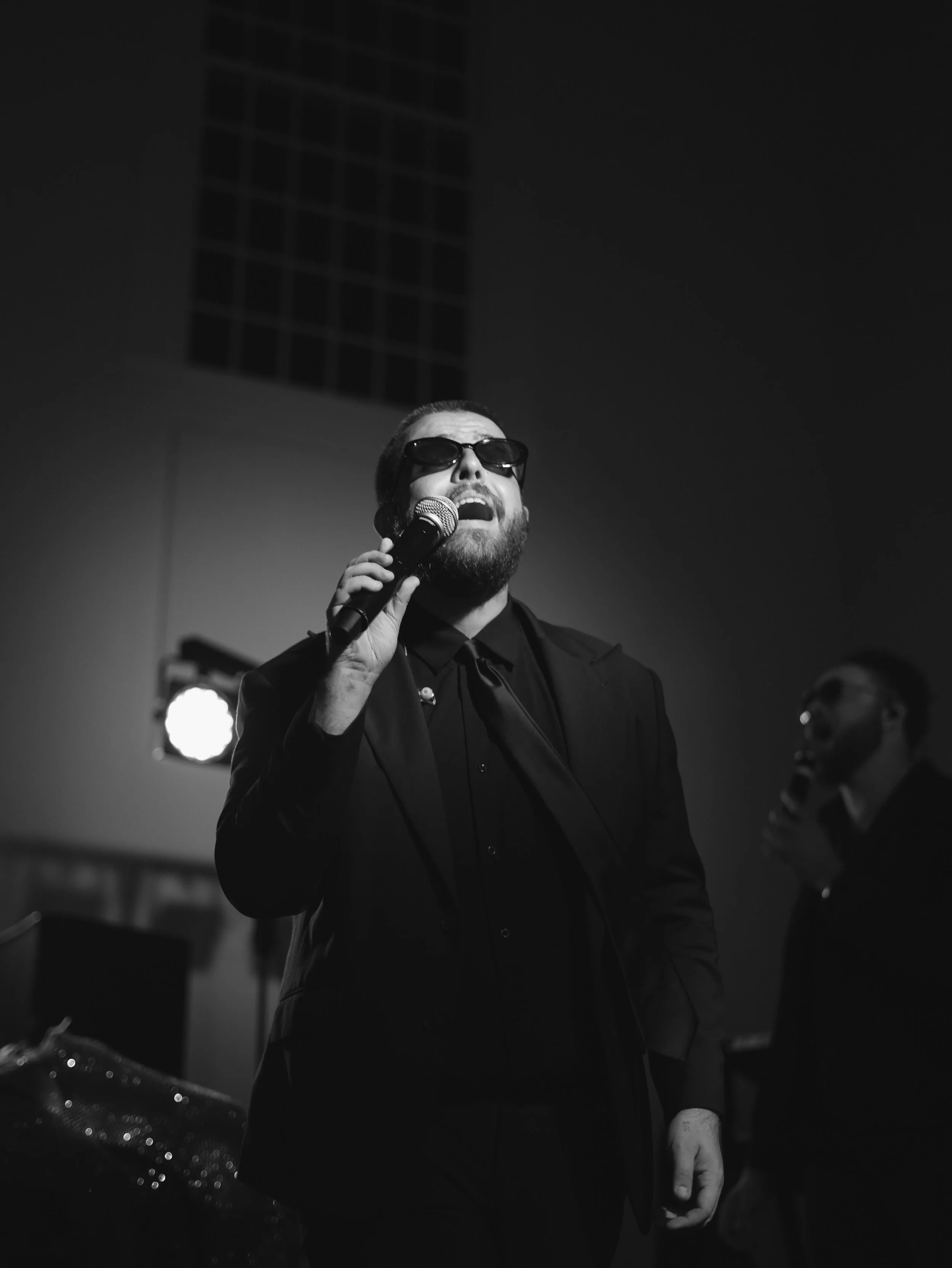 A man with a beard wearing sunglasses singing into a microphone with a woman in the background, in a black and white photo.
