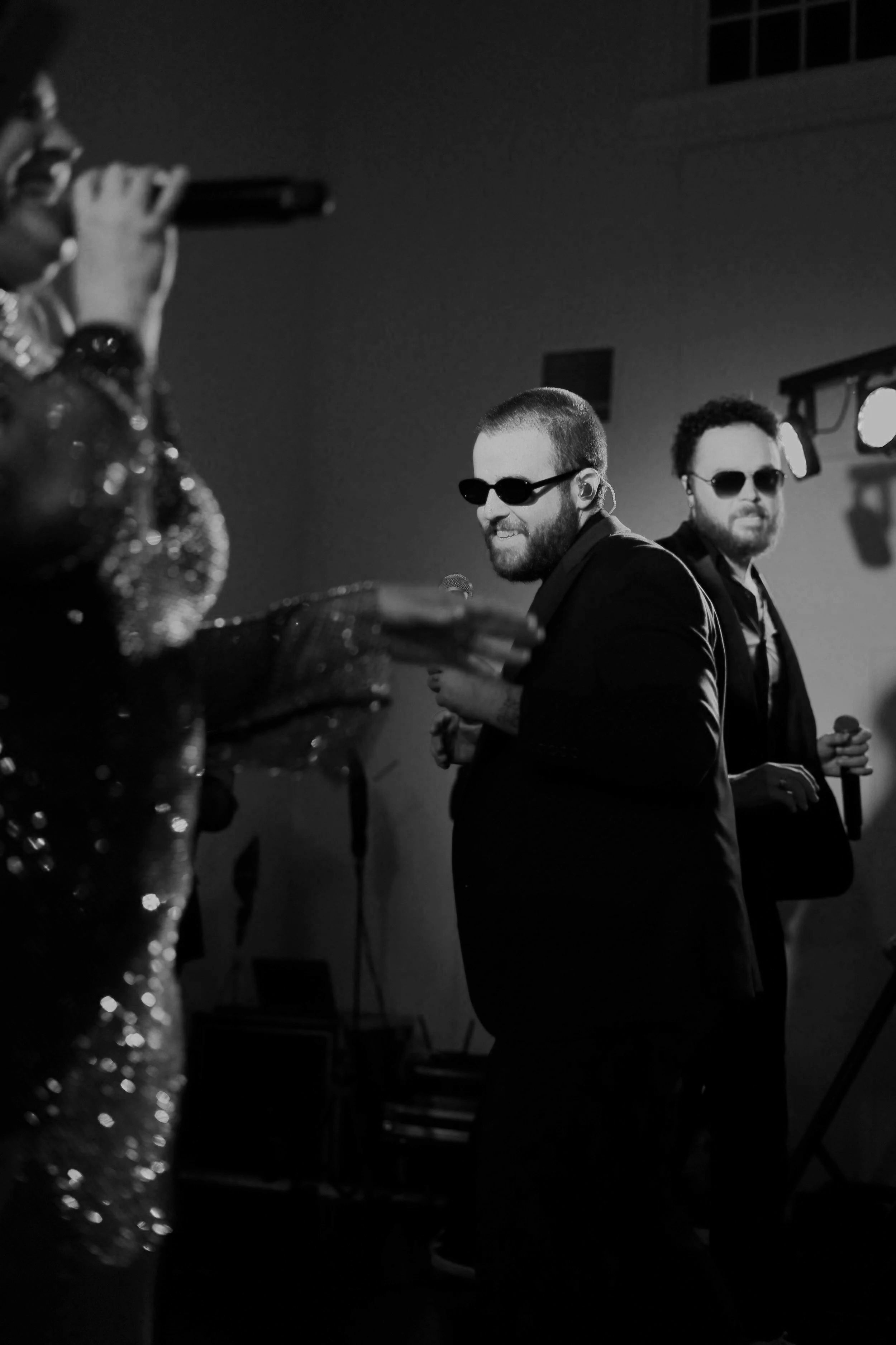 Black and white photo of two men with sunglasses performing on stage, with a partially visible singer in the foreground.