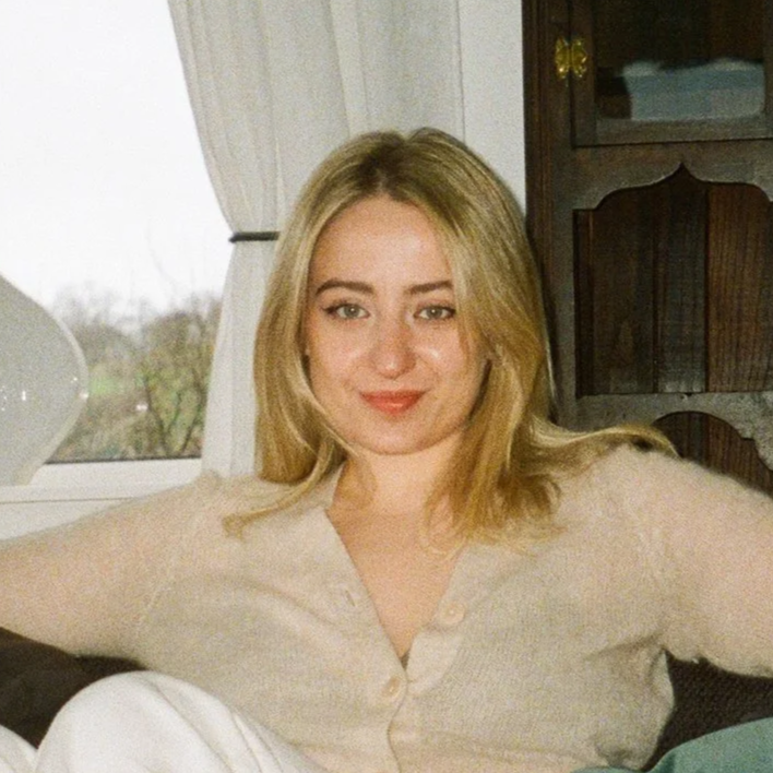 A young woman with blonde hair sitting comfortably indoors on a sofa, smiling at the camera, with a window and curtains in the background.