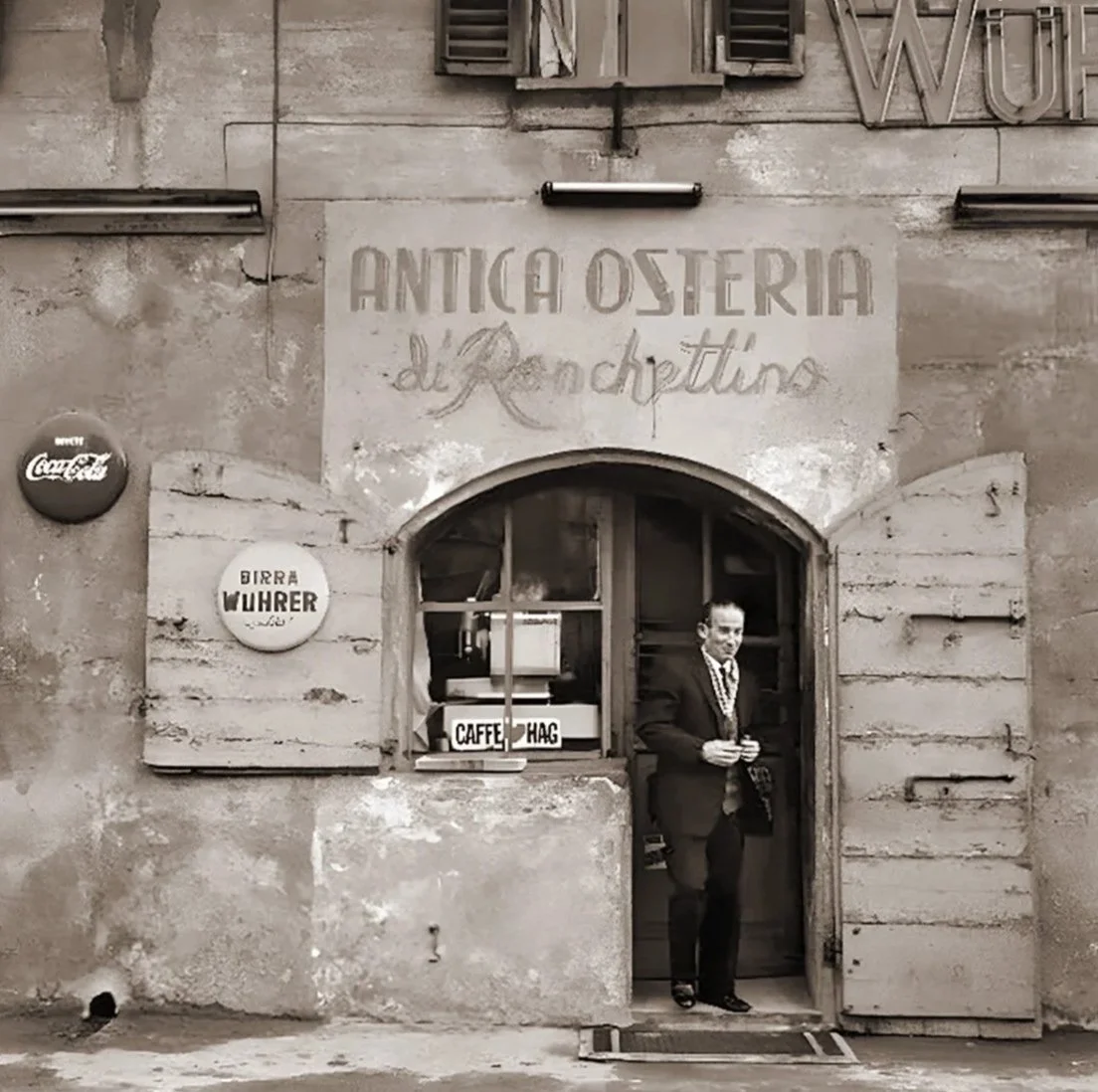 Esterno di una vecchia osteria in stile rustico con un uomo in abito elegante che esce, sul muro ci sono cartelli pubblicitari e una scritta che recita 'Antica Osteria di Rachittino'.