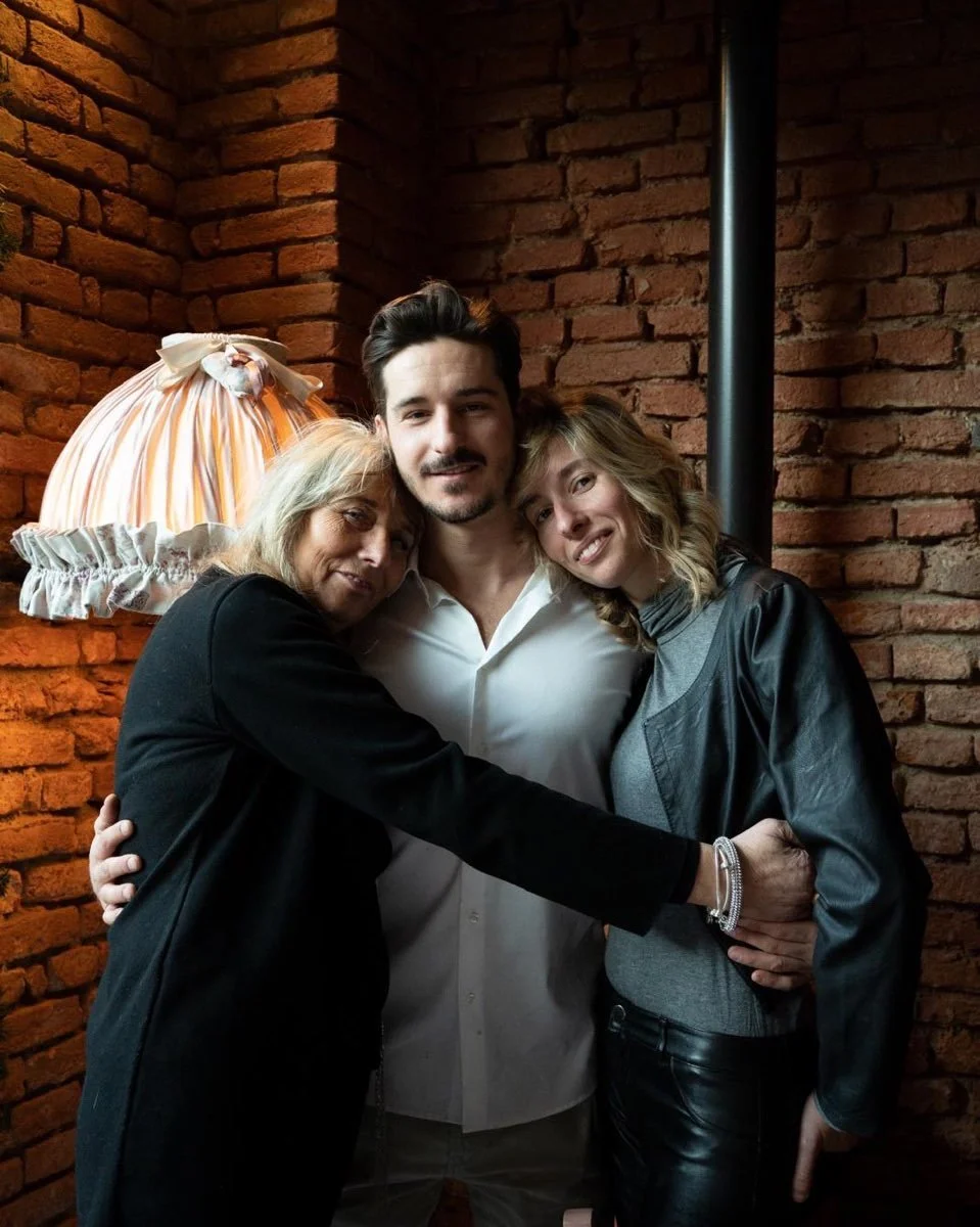 Tre persone si abbracciano tra loro davanti a una parete di mattoni rossi, con una lampada con paralume a righe sullo sfondo.
