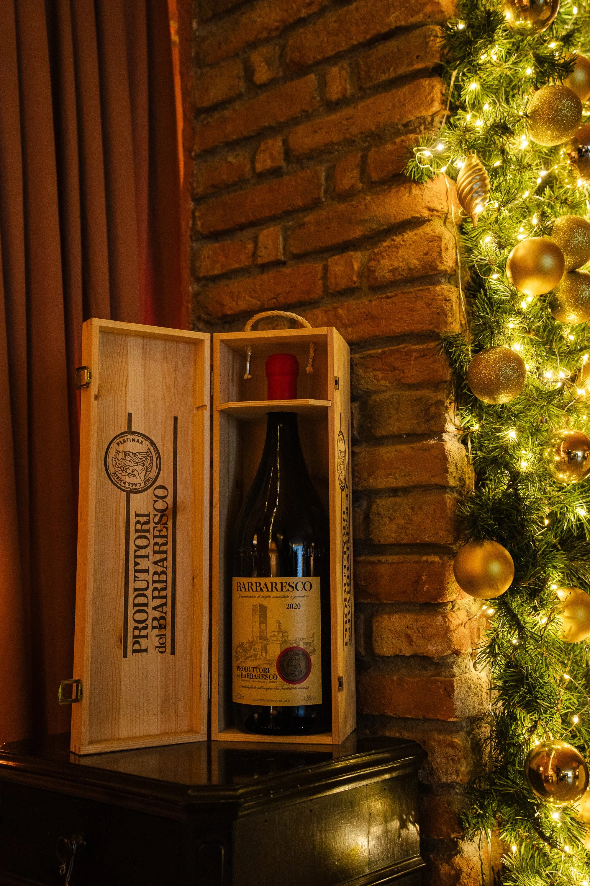 Una bottiglia di vino all'interno di una scatola di legno vicino a un camino di mattoni decorato con luci natalizie e palline dorate.