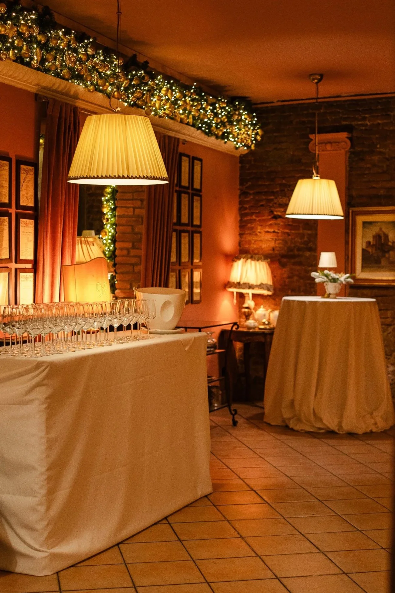Interno di una sala decorata per le feste natalizie con luci e ghirlande, tavoli con tovaglia bianca, lampade e quadri alle pareti.