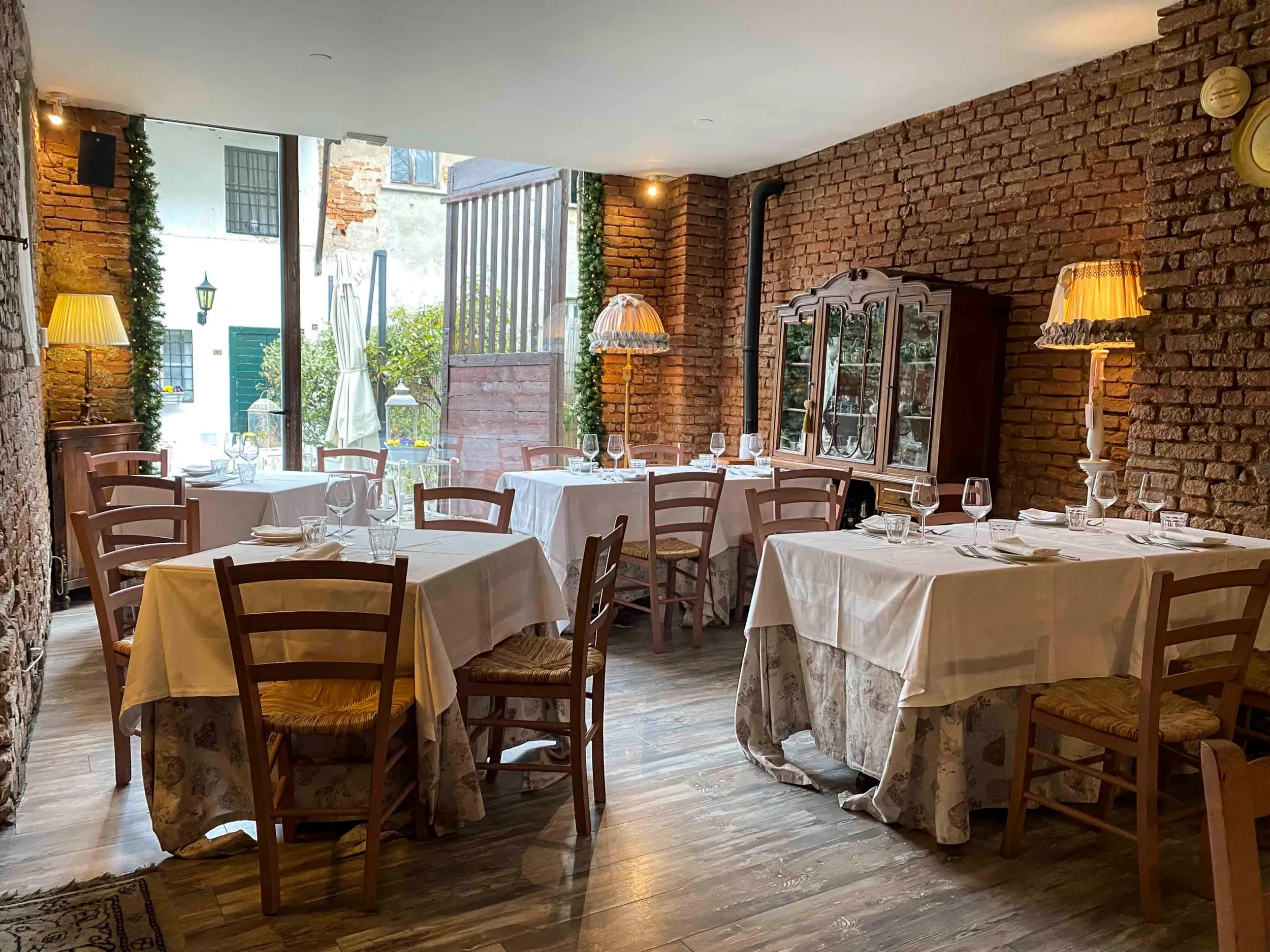 Sala da pranzo elegante con pareti di mattoni a vista, tavoli con tovaglie bianche, bicchieri e posate, sedie di legno, lampade da tavolo e naturale illuminazione dall'esterno.