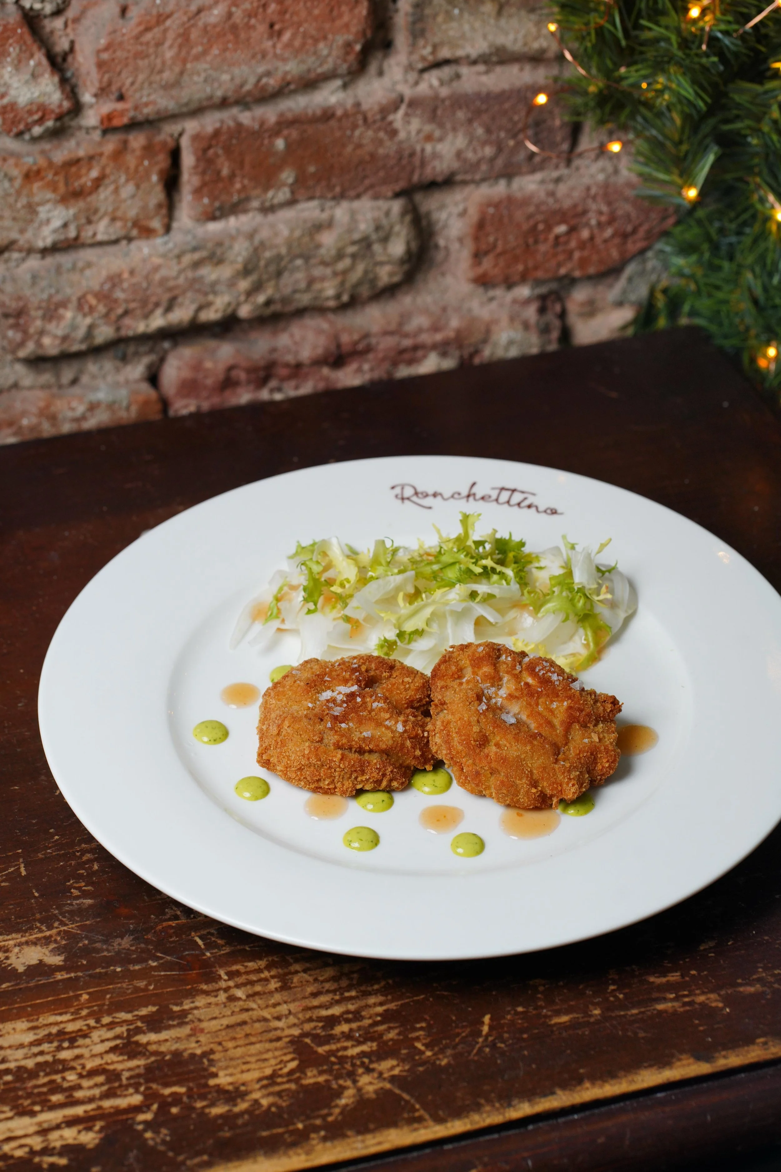 Piatti di pollo impanato con insalata, serviti su un piatto bianco con scritta 'Rocchettino', su una tavola di legno, con parete in mattoni sullo sfondo.