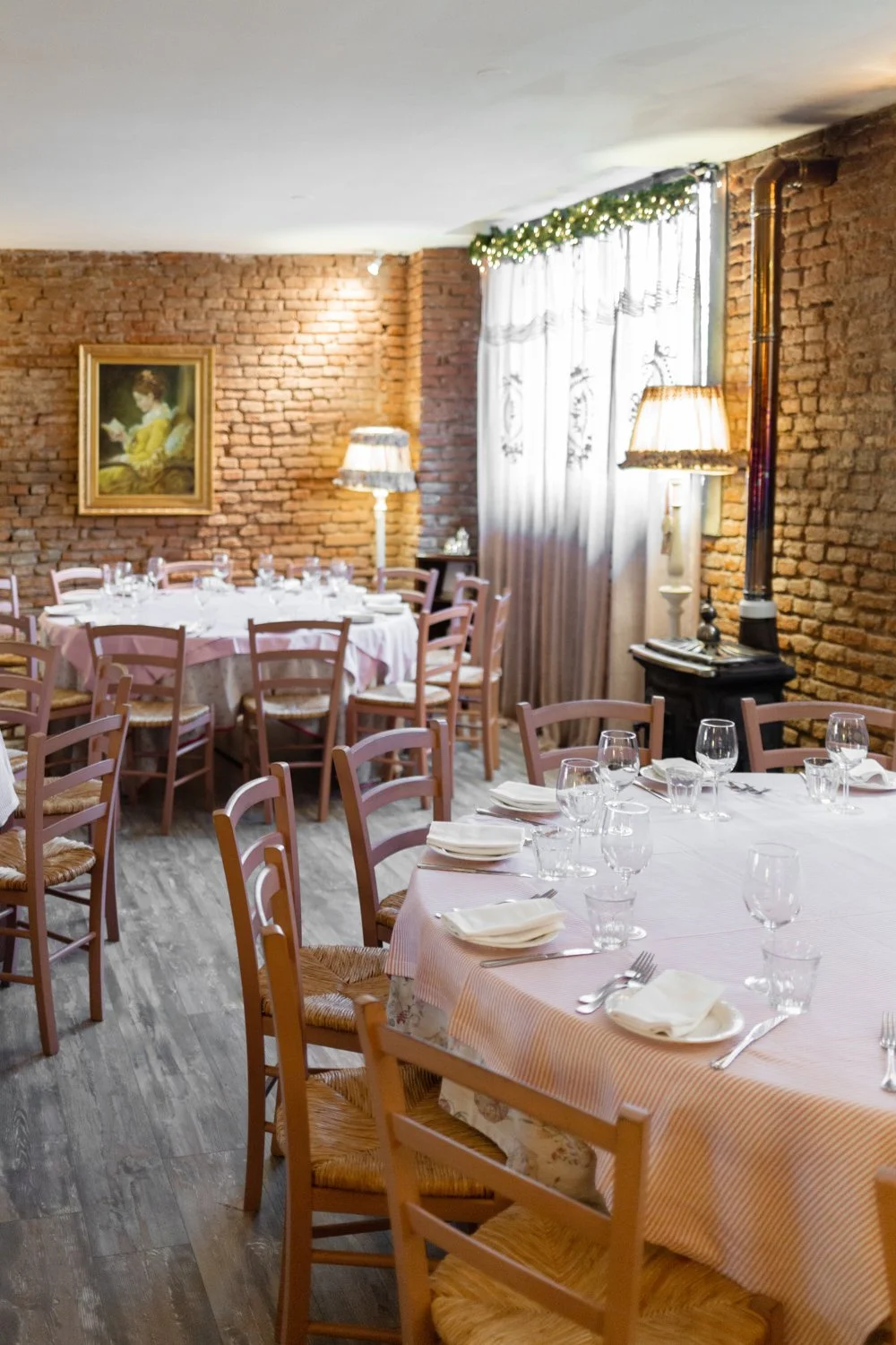 Interno di ristorante elegante con tavoli apparecchiati, pareti di mattoni, quadri, candele e decorazioni natalizie.