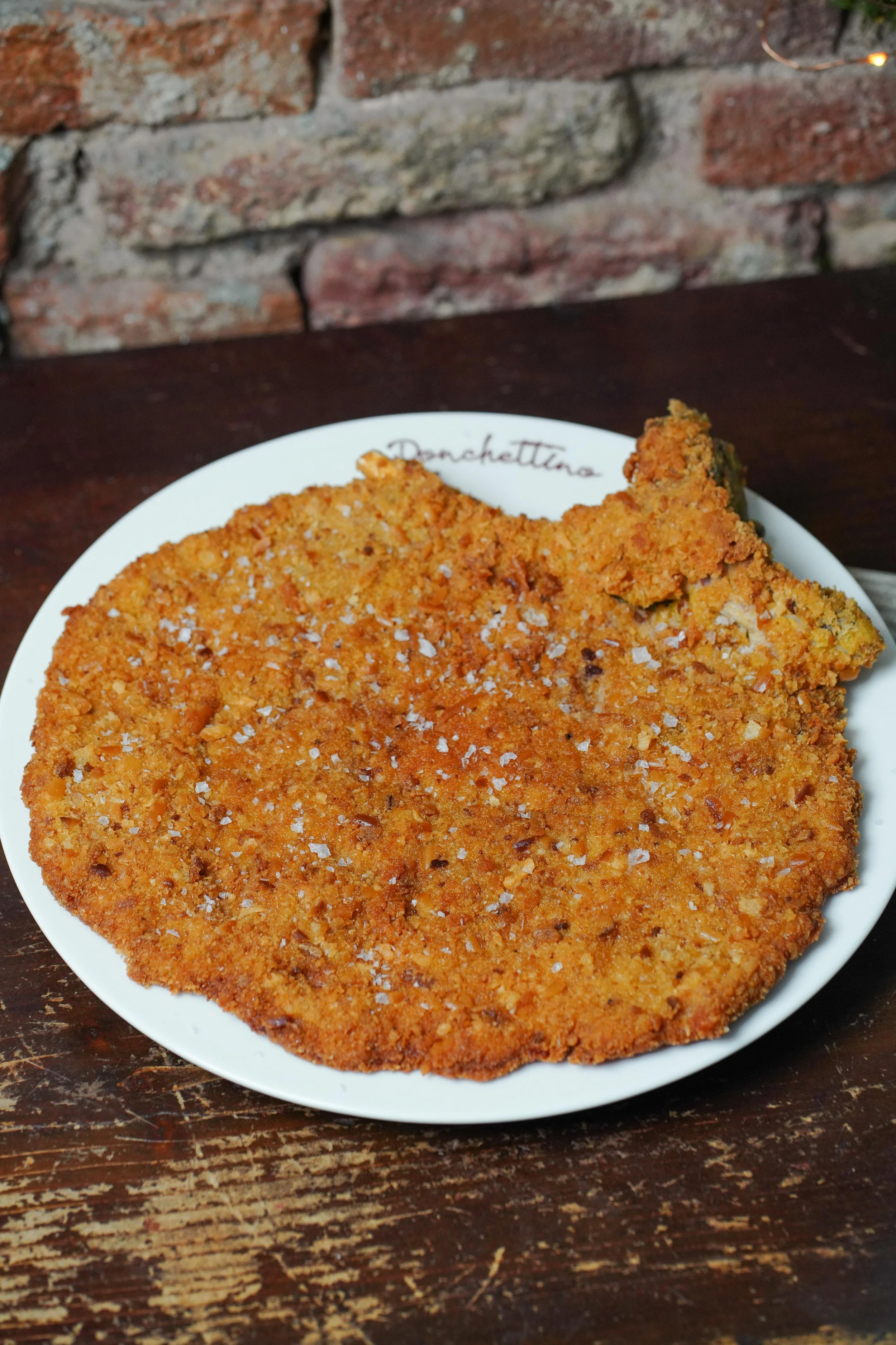 Cotoletta alla milanese su piatto bianco con scritto 'Donchettina', posata su tavolo di legno con sfondo di muro di mattoni.