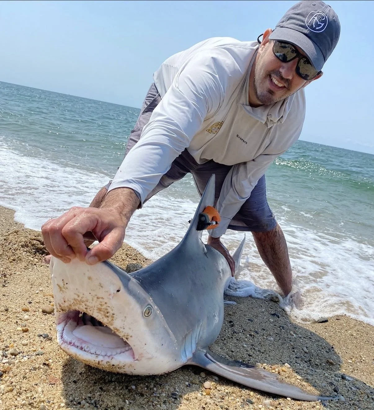 A man on a beach holding a large shark with a wide-open mouth. The man is wearing sunglasses, a cap, and light-colored clothing. The ocean and clear sky are visible in the background.