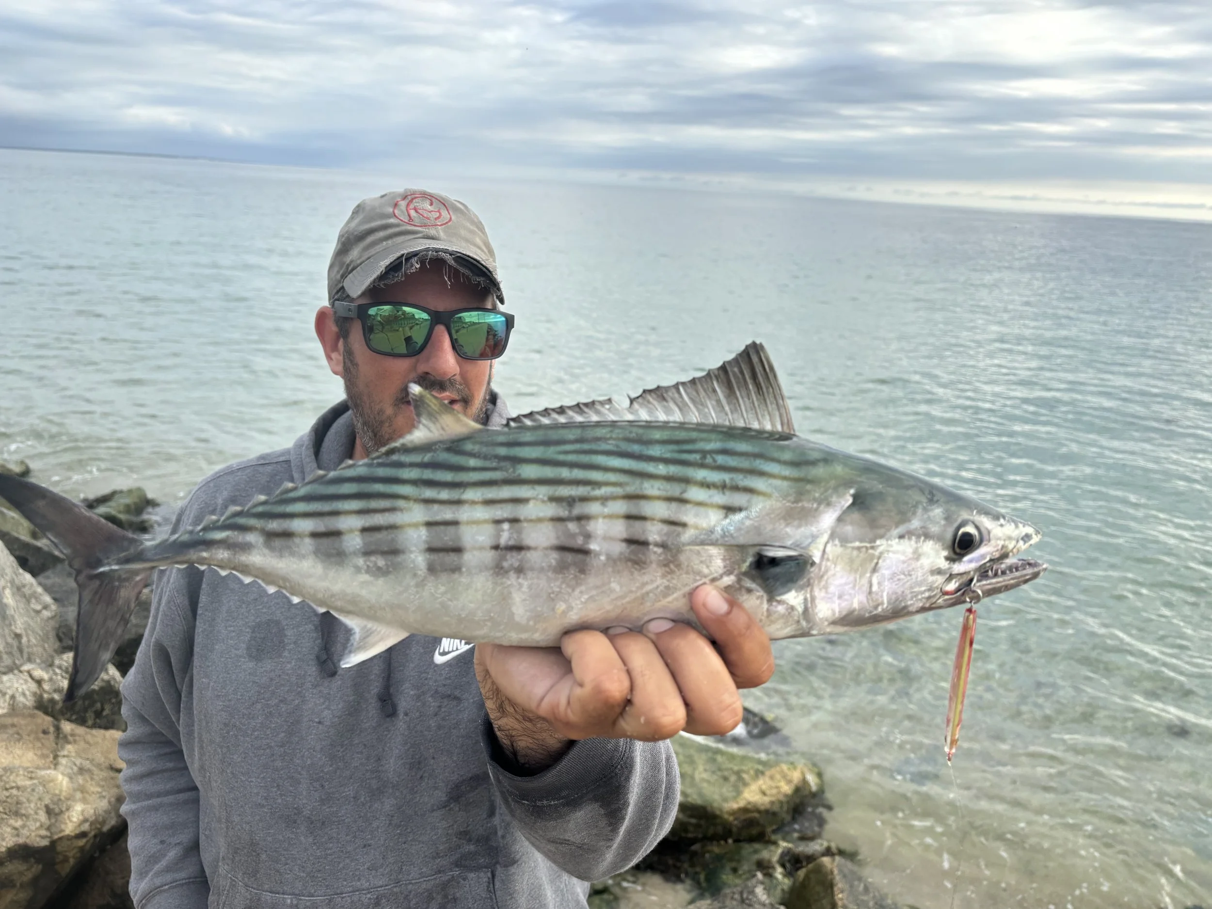 Man wearing a gray hoodie, sunglasses, and a baseball cap holding a large fish near the shore with water and rocks in the background.