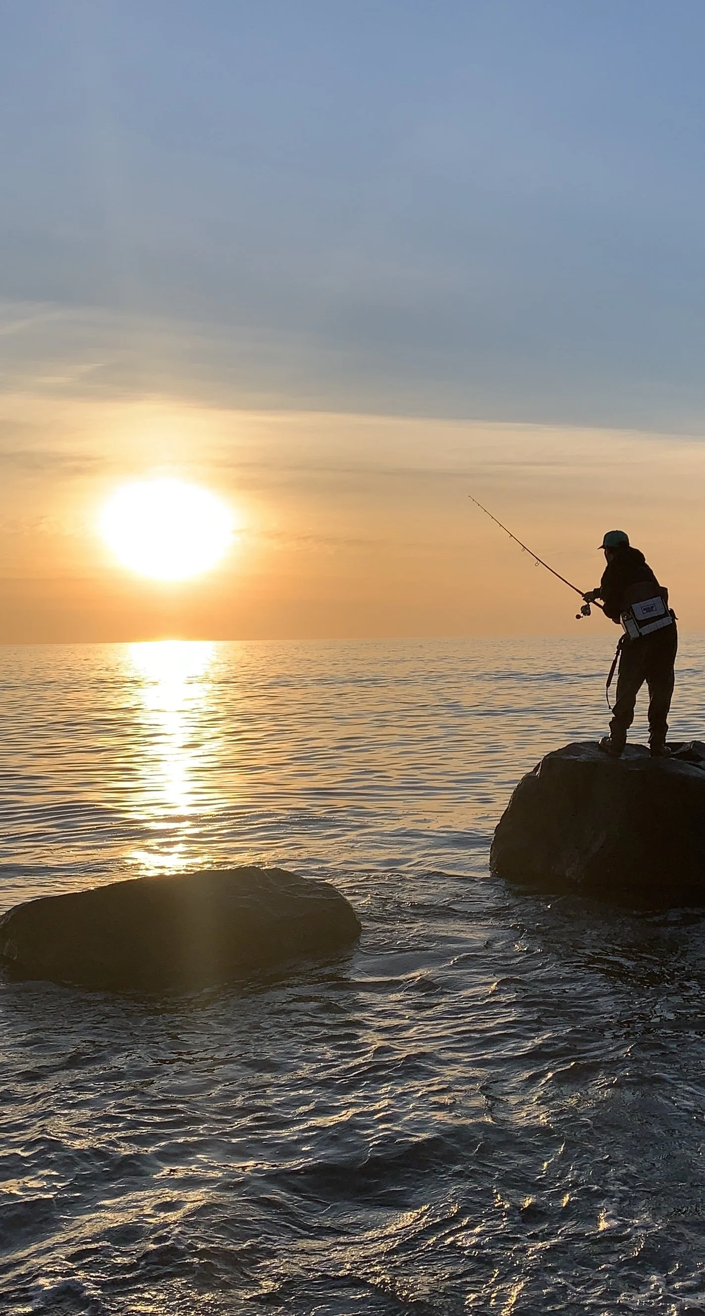 A person fishing on rocks at sunset over the ocean.