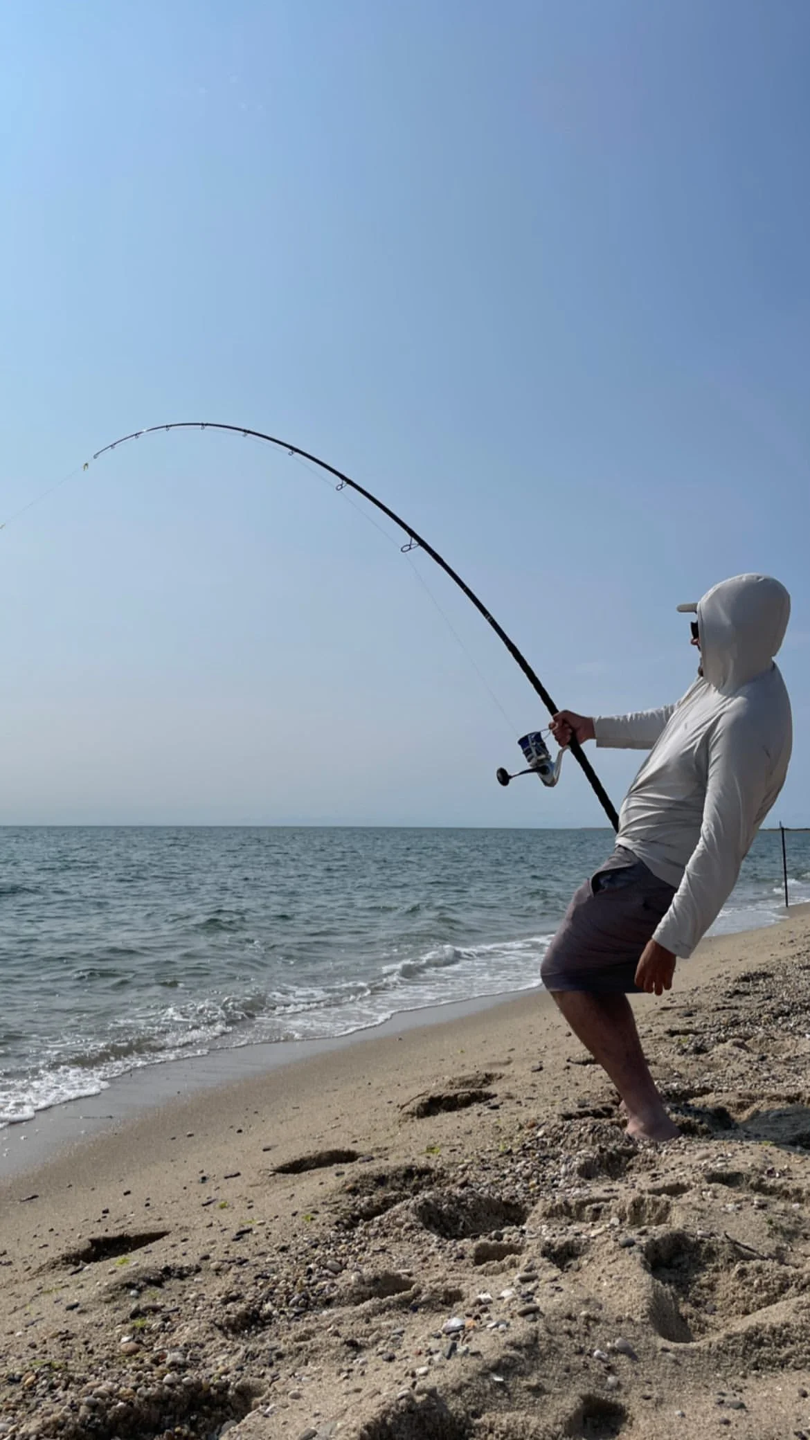 Person fishing on a beach, wearing a hoodie and shorts, holding a fishing rod over the water.