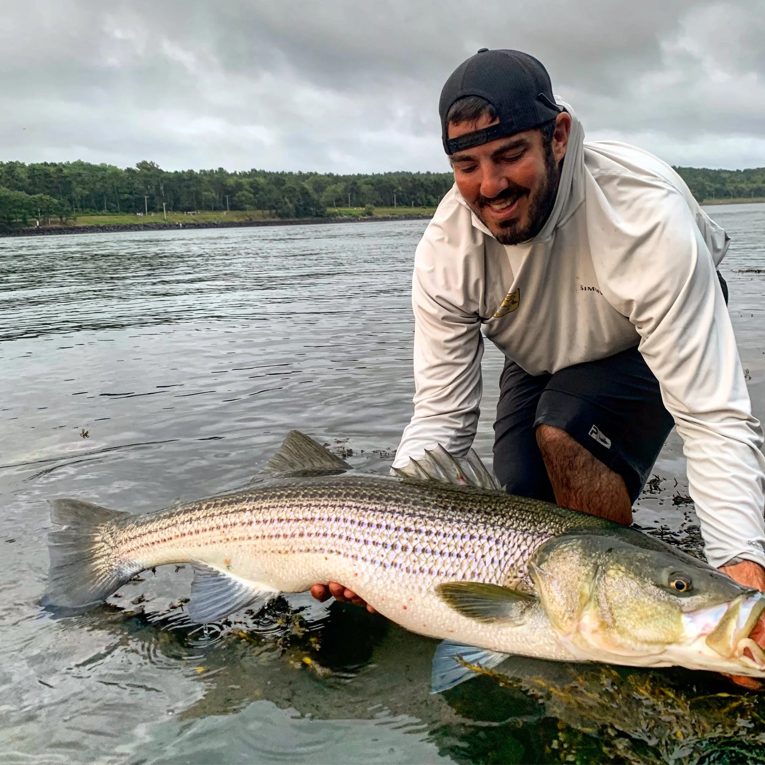 Man kneeling in water, holding a large fish, smiling with a cloudy sky and trees in the background.