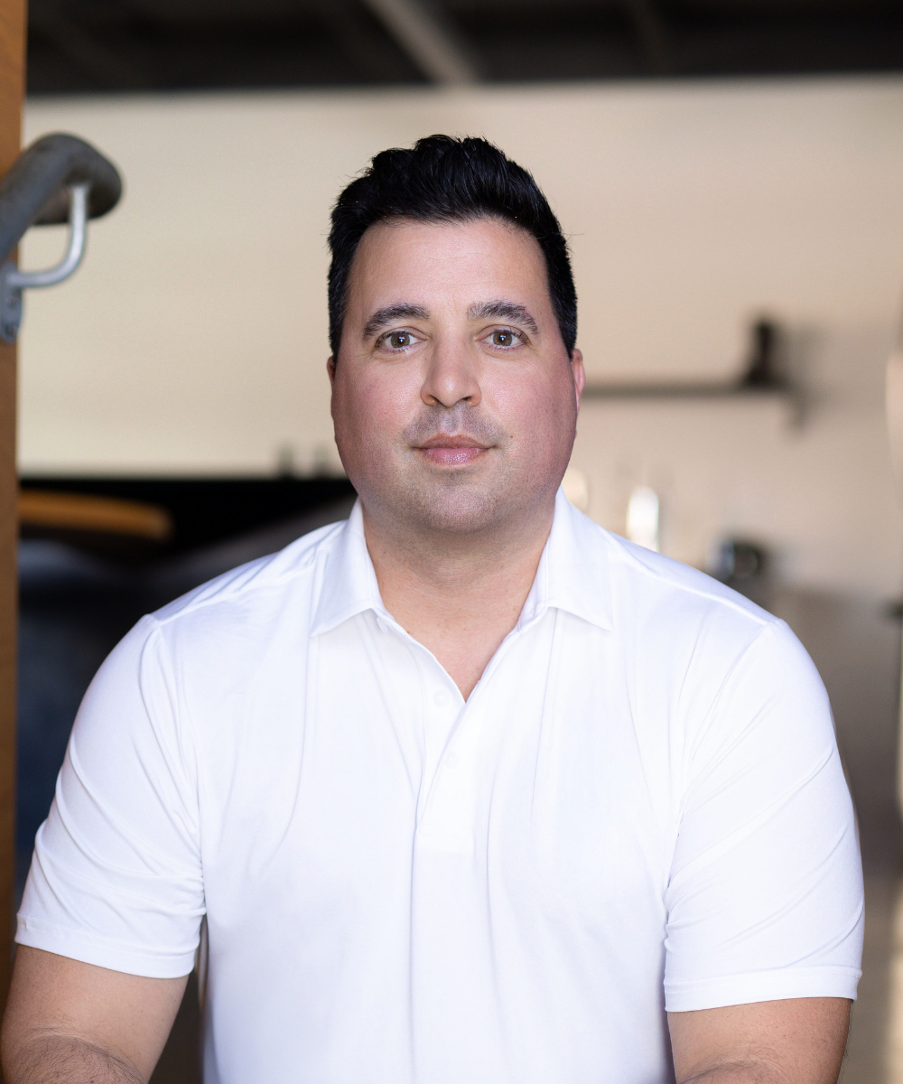A man with dark hair and light skin wearing a white polo shirt, sitting indoors with a blurred background.