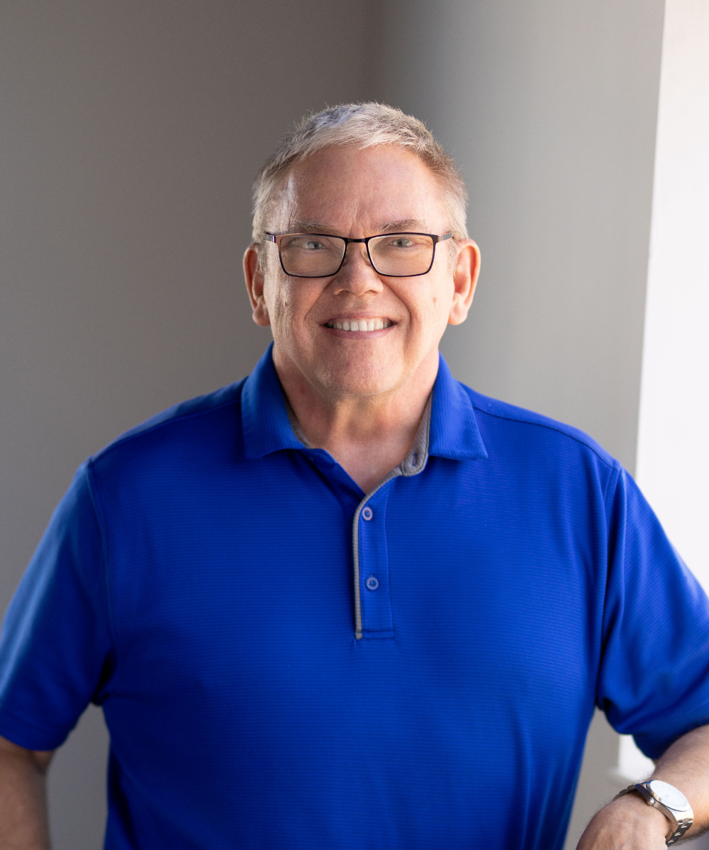 A smiling middle-aged man with glasses, wearing a blue polo shirt, standing indoors near a window with natural light.
