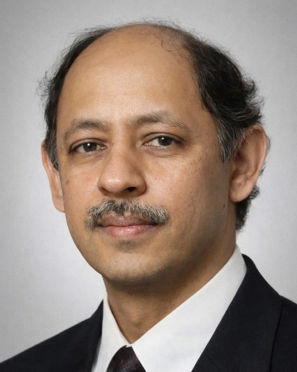 Professional headshot of a man with light brown skin, short black hair, and a mustache, wearing a black suit and white shirt, against a plain light gray background.