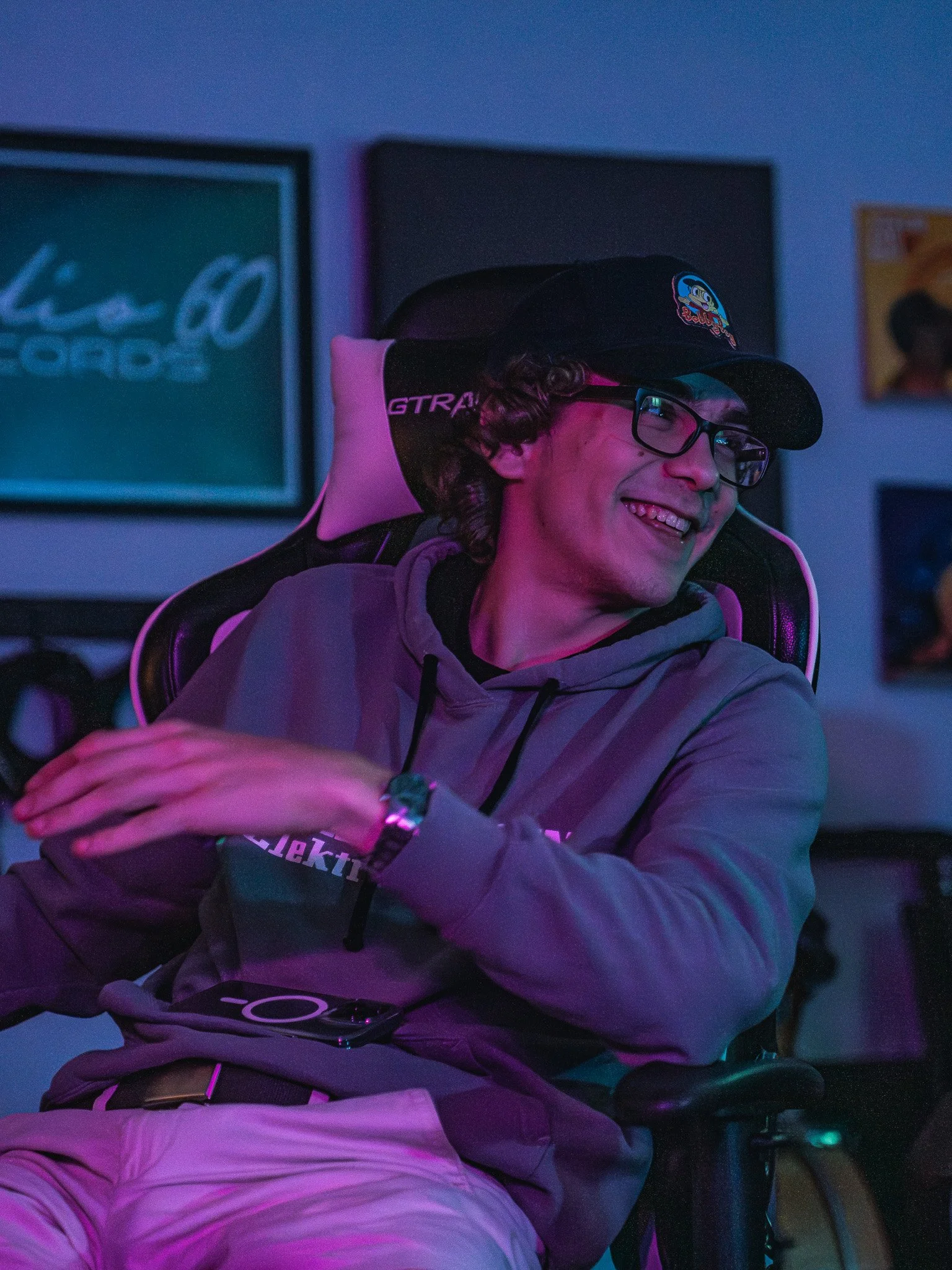 Young man with glasses and curly hair smiling, sitting in a gaming chair in a dimly lit room with colorful lights and framed posters on the wall.