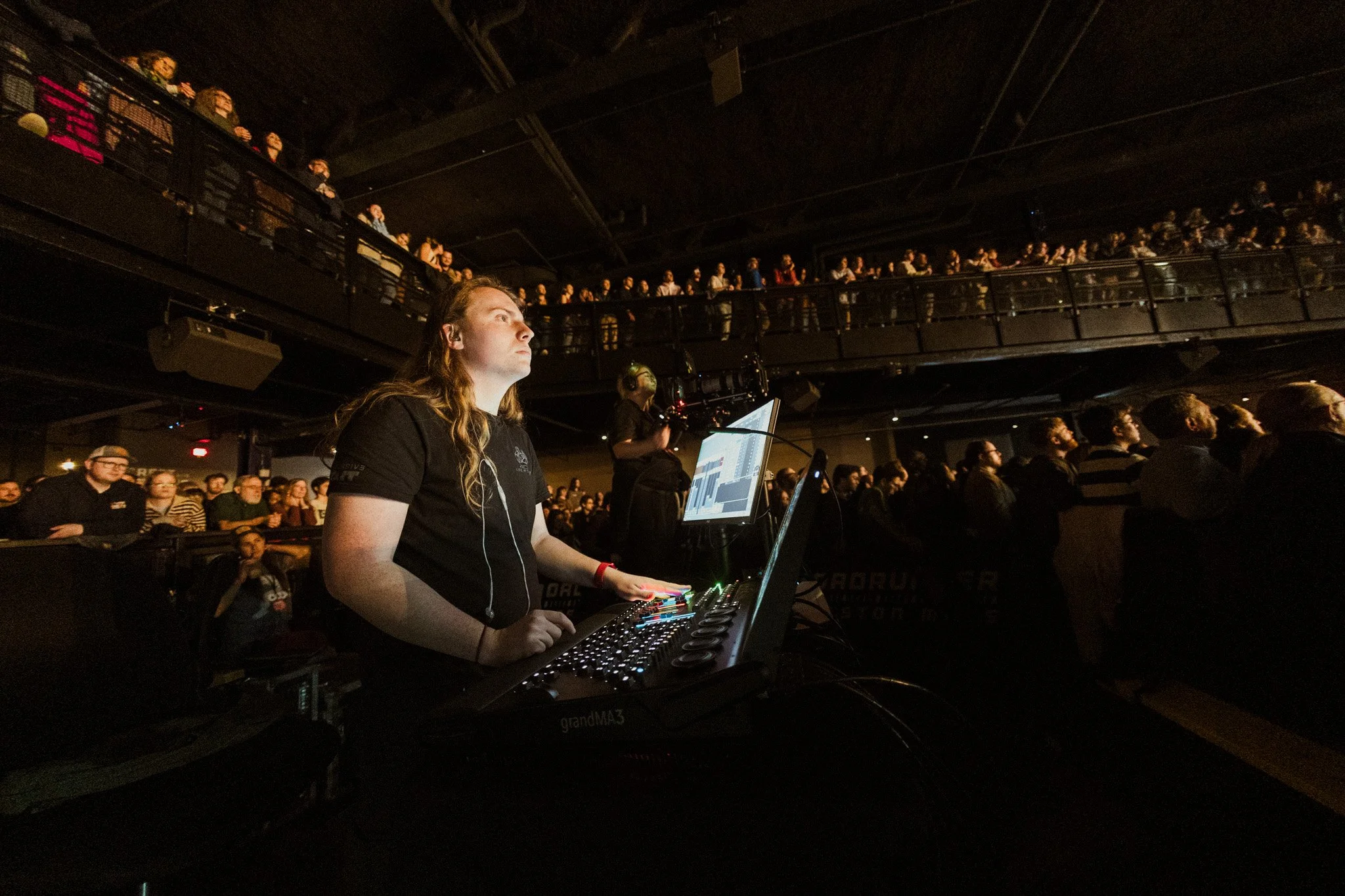 A female DJ with long hair, wearing a black shirt, stands behind a DJ setup with a laptop and audio equipment, lighting and people in a dark concert venue with a balcony filled with audience members.