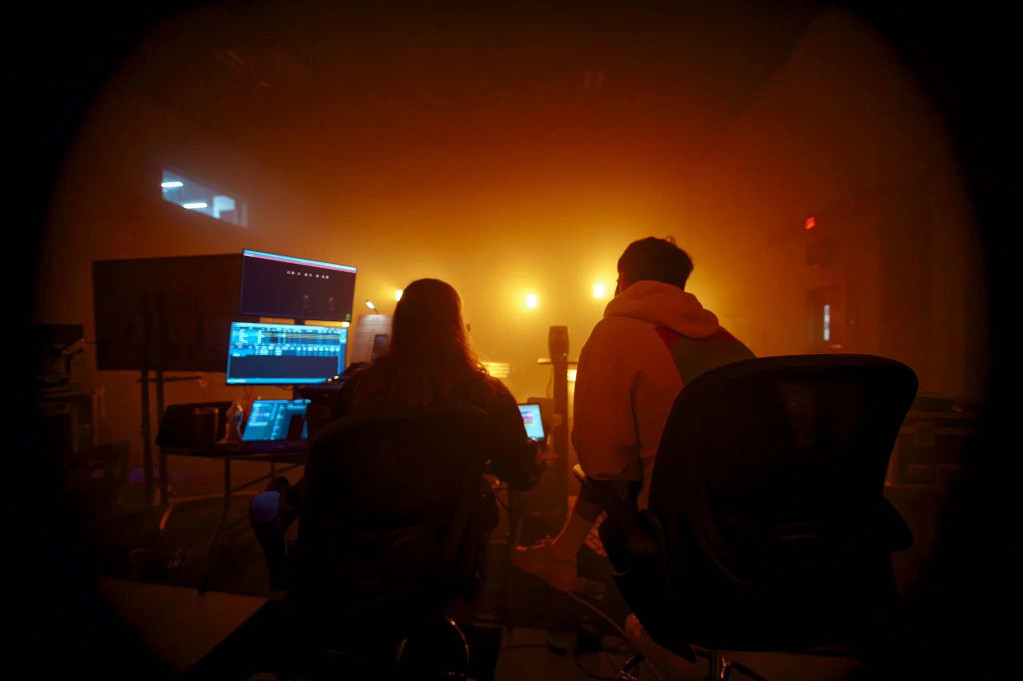Two people sitting at a computer setup in a dimly lit room with colorful lighting, possibly a nightclub or a music production studio.