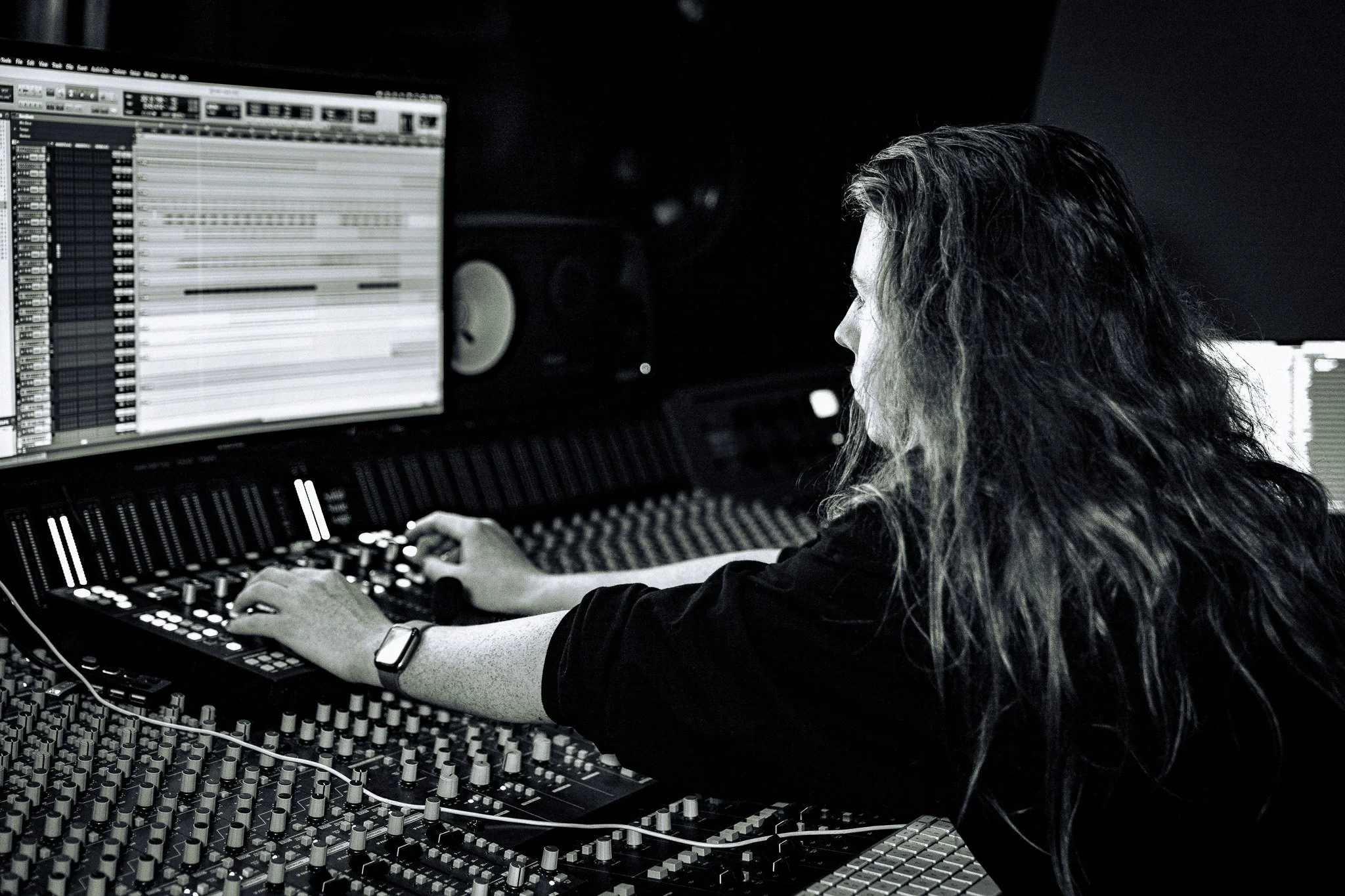 A woman with long, wavy hair working at a music mixing console in a dark studio with computer screens displaying digital audio workstations.