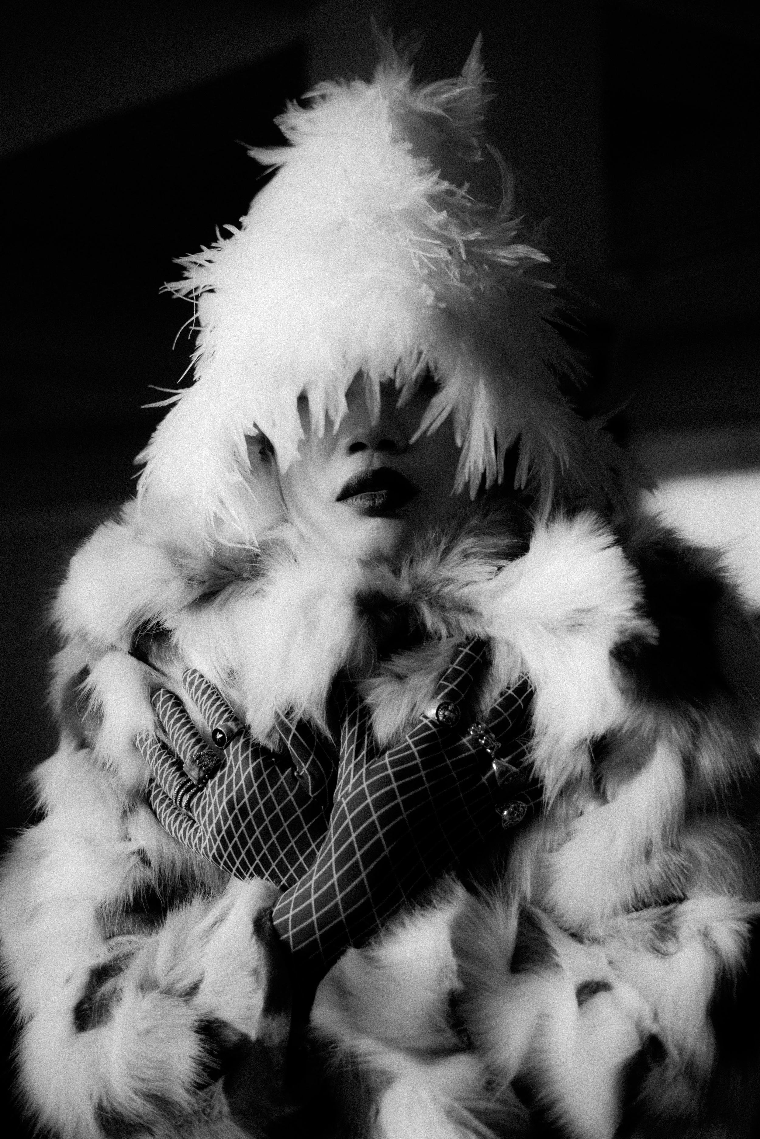 A person wearing a large, fluffy, feathered hat and matching coat, with dark lipstick and gloves with a geometric pattern, covering one eye with the hat.