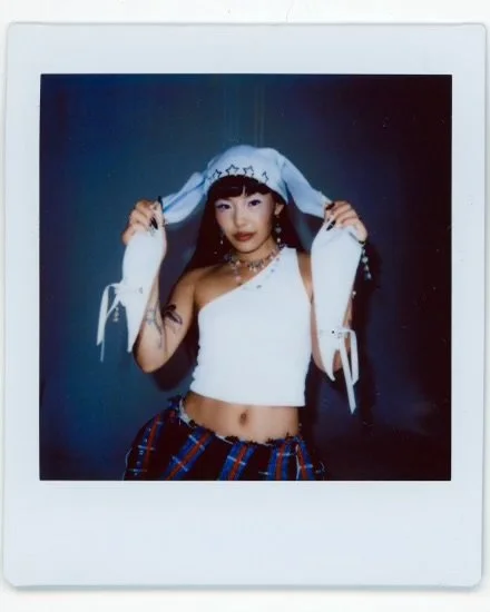 Young woman with dark hair, wearing a white cropped top, plaid pants, and a white hoodie with star patterns, posing against a dark background.