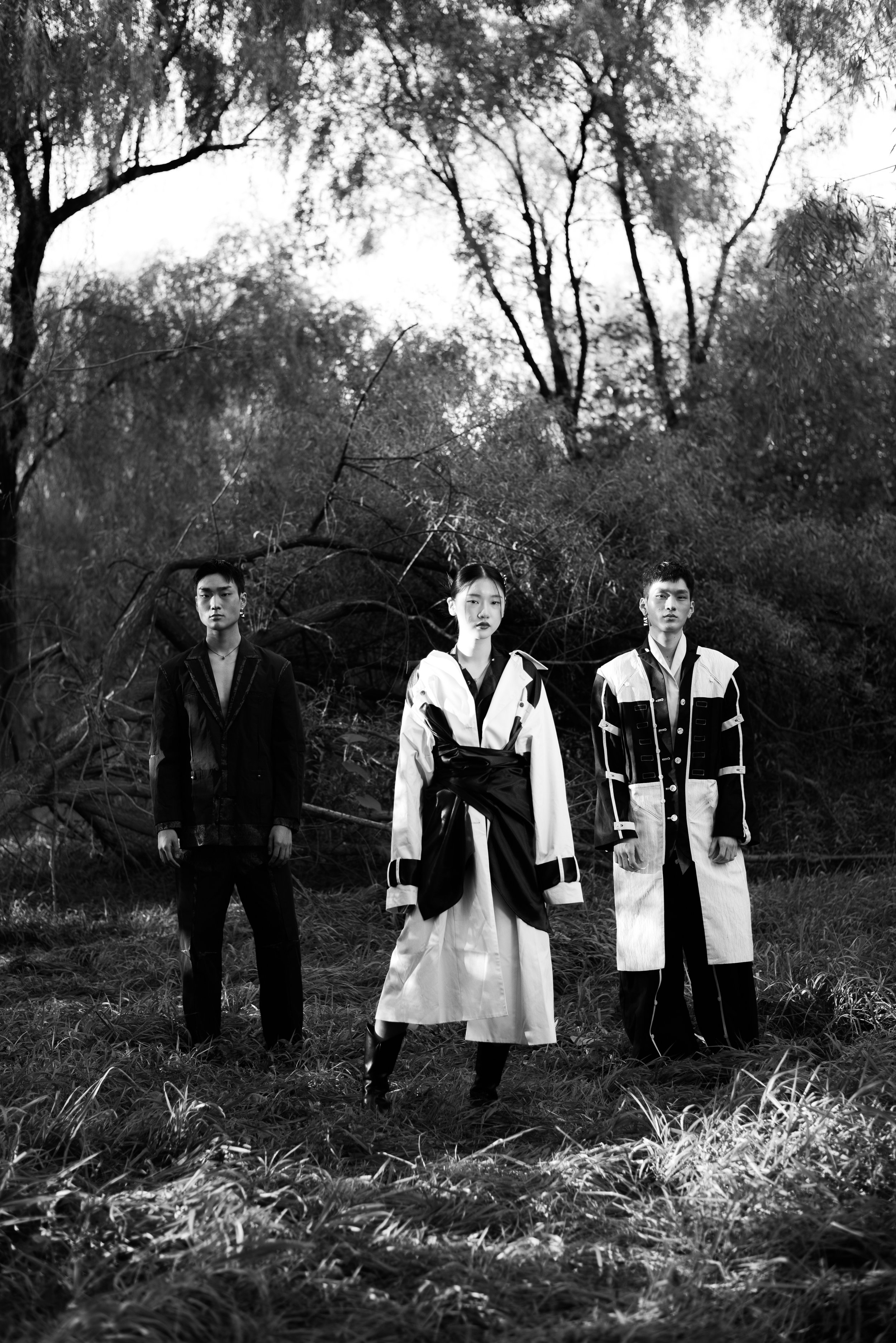 Three individuals standing in a grassy field with trees and foliage in the background, all dressed in something resembling futuristic or avant-garde fashion in black and white.